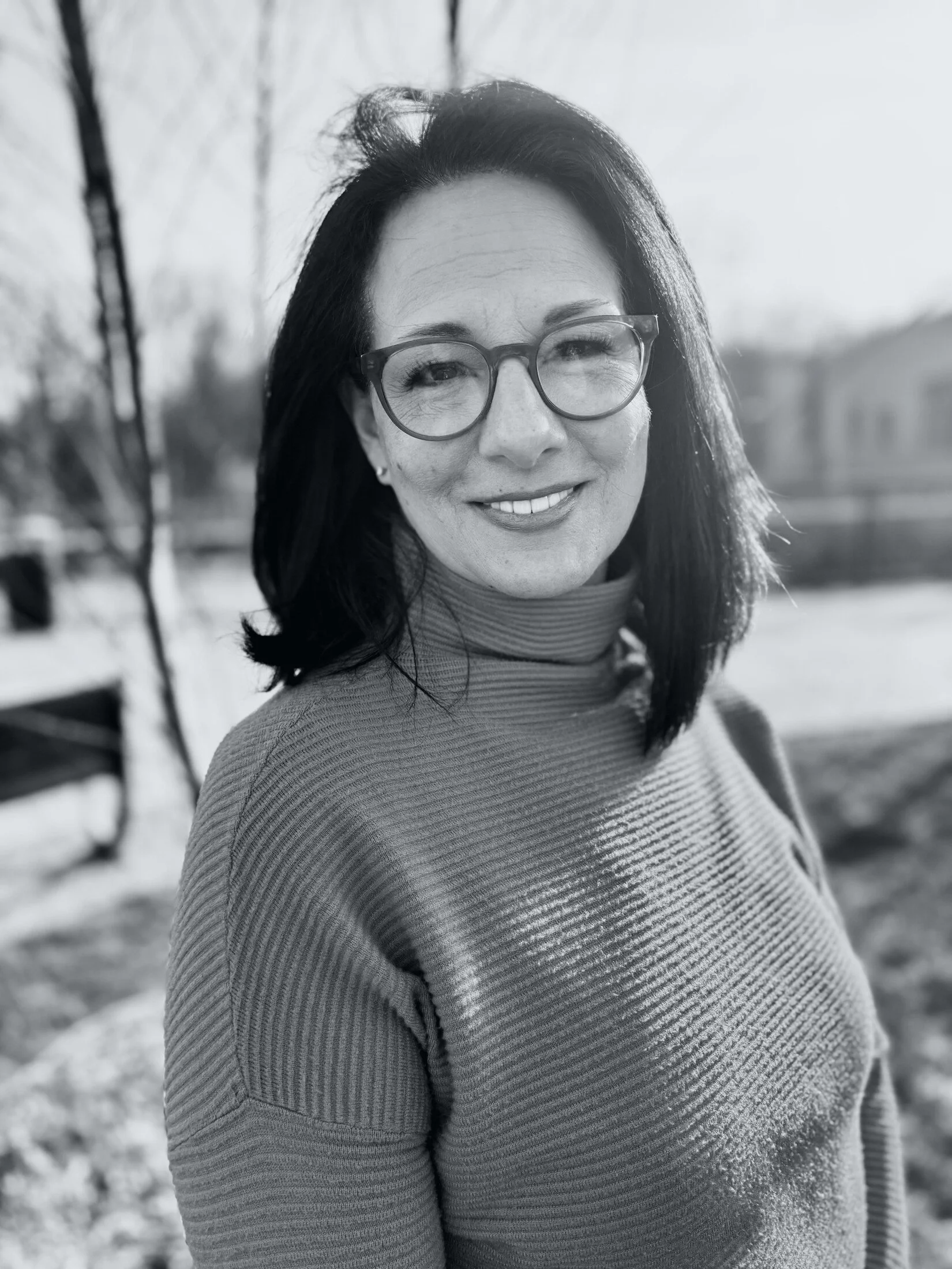 A woman with dark hair and glasses smiling outdoors in a black and white photo, with trees and a bench in the background.