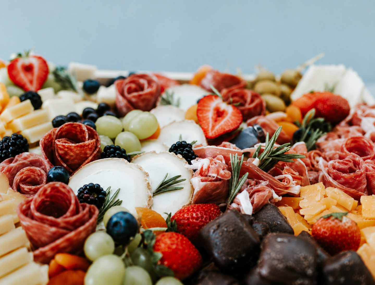 Close-up of a charcuterie platter with cheese, berries, strawberries, grapes, cured meats, chocolates, and herbs against a blurred light blue background.