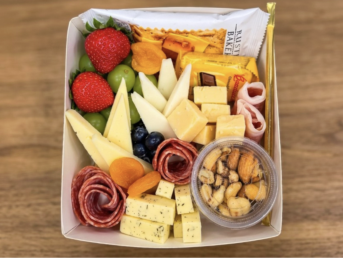 A cheese and fruit platter with strawberries, green grapes, blueberries, dried apricots, assorted cheeses, sliced meats, mixed nuts, a chocolate bar, and a cracker