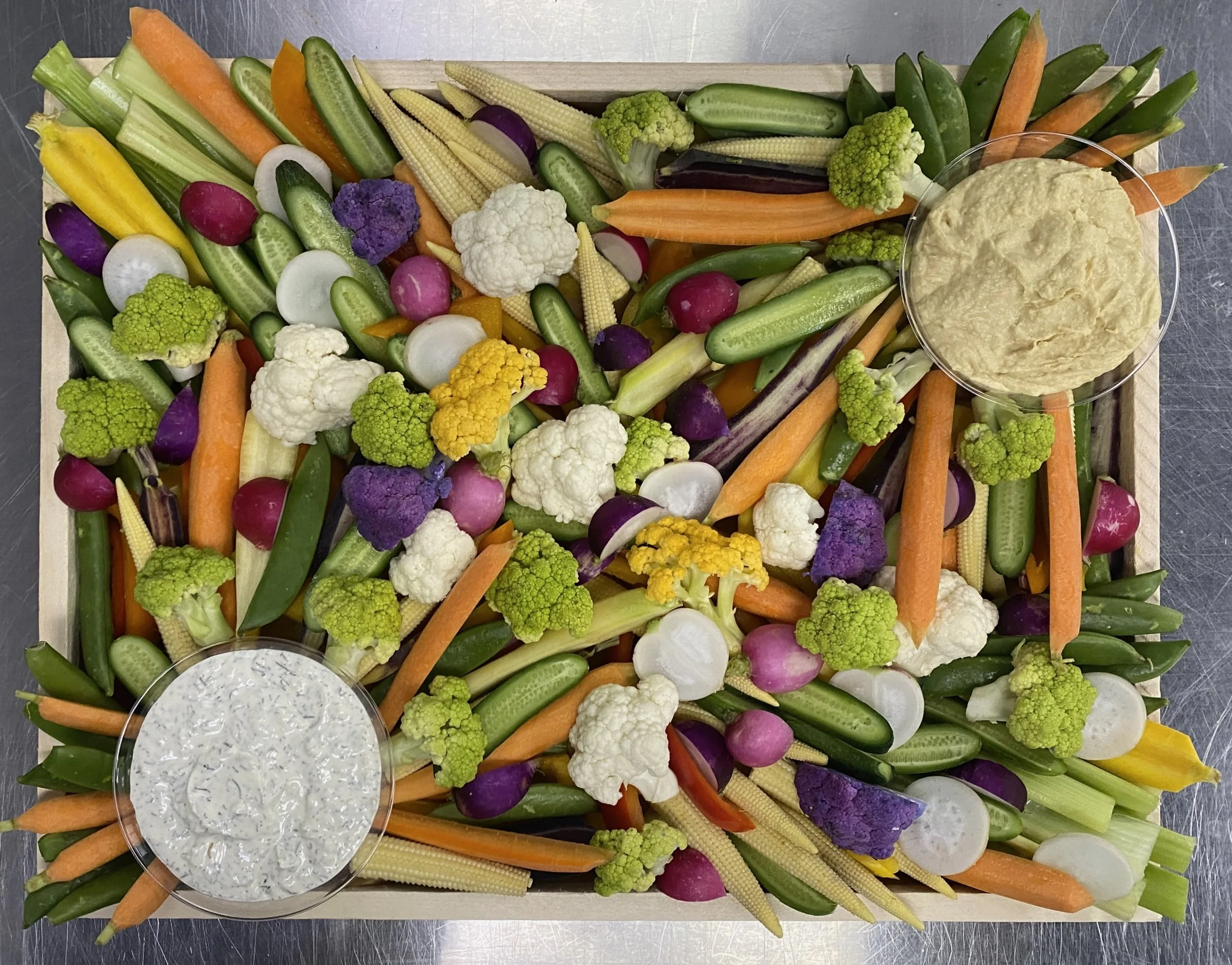 Assorted fresh vegetables including carrots, radishes, cauliflower, broccoli, cucumbers, baby corn, and yellow squash, with a bowl of creamy dip on a wooden tray.