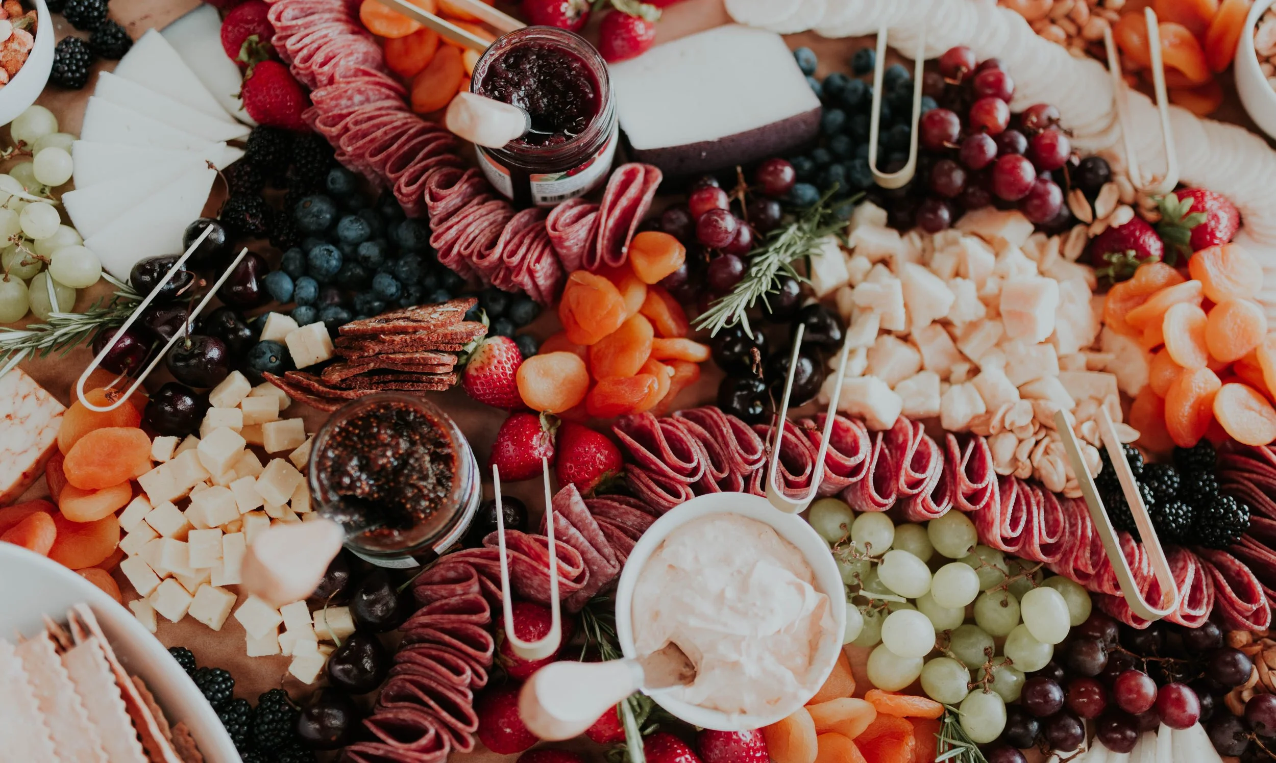 A close-up view of a cheese and fruit platter. It features assorted cheeses, grapes, berries, sliced apricots, and various meats and crackers, arranged for a festive gathering.