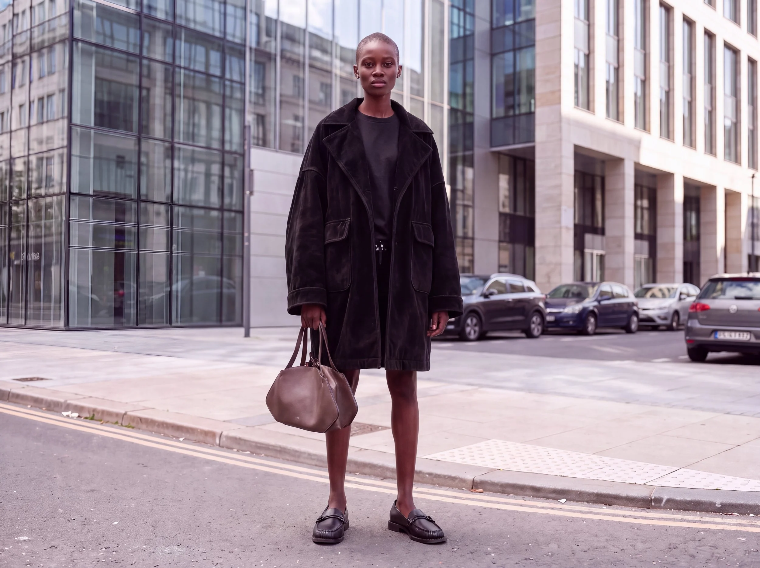 Donna con abbigliamento nero, ma senza scarpe, che cammina su un marciapiede di fronte a edifici moderni in città.