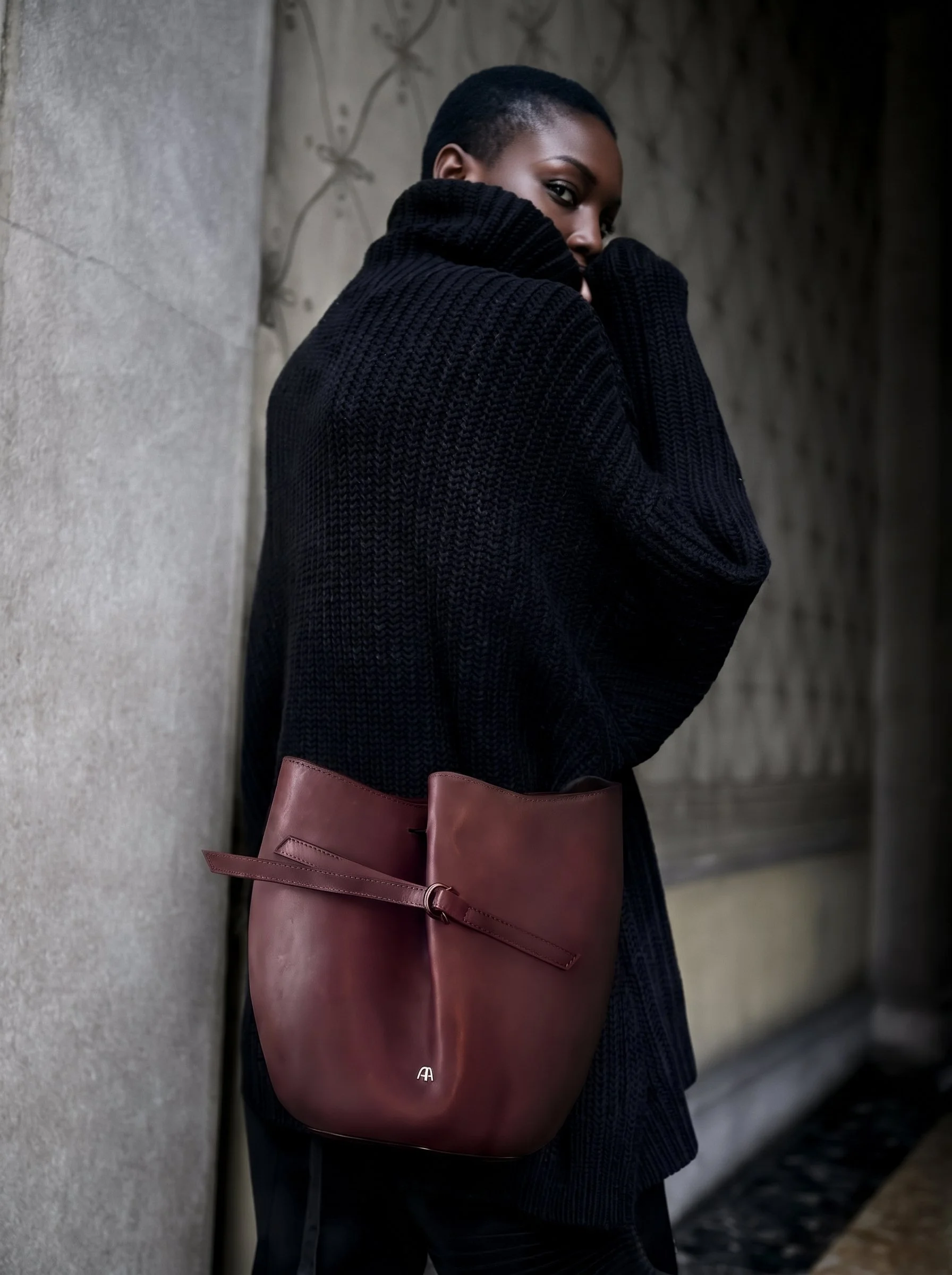Ragazza con felpa nera che si nasconde il volto, portando una borsa marrone, in un ambiente urbano con pareti di cemento.