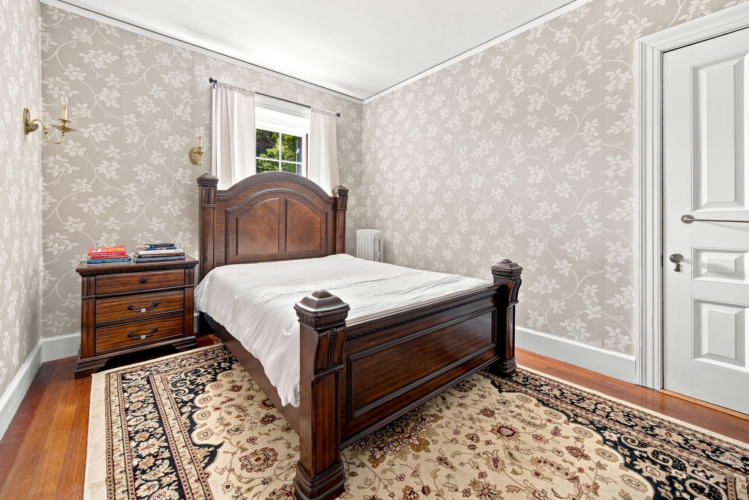 A bedroom with a wooden bed frame, a nightstand with books, a window with white curtains, a radiator, patterned area rug, beige floral wallpaper, and white ceiling and door.