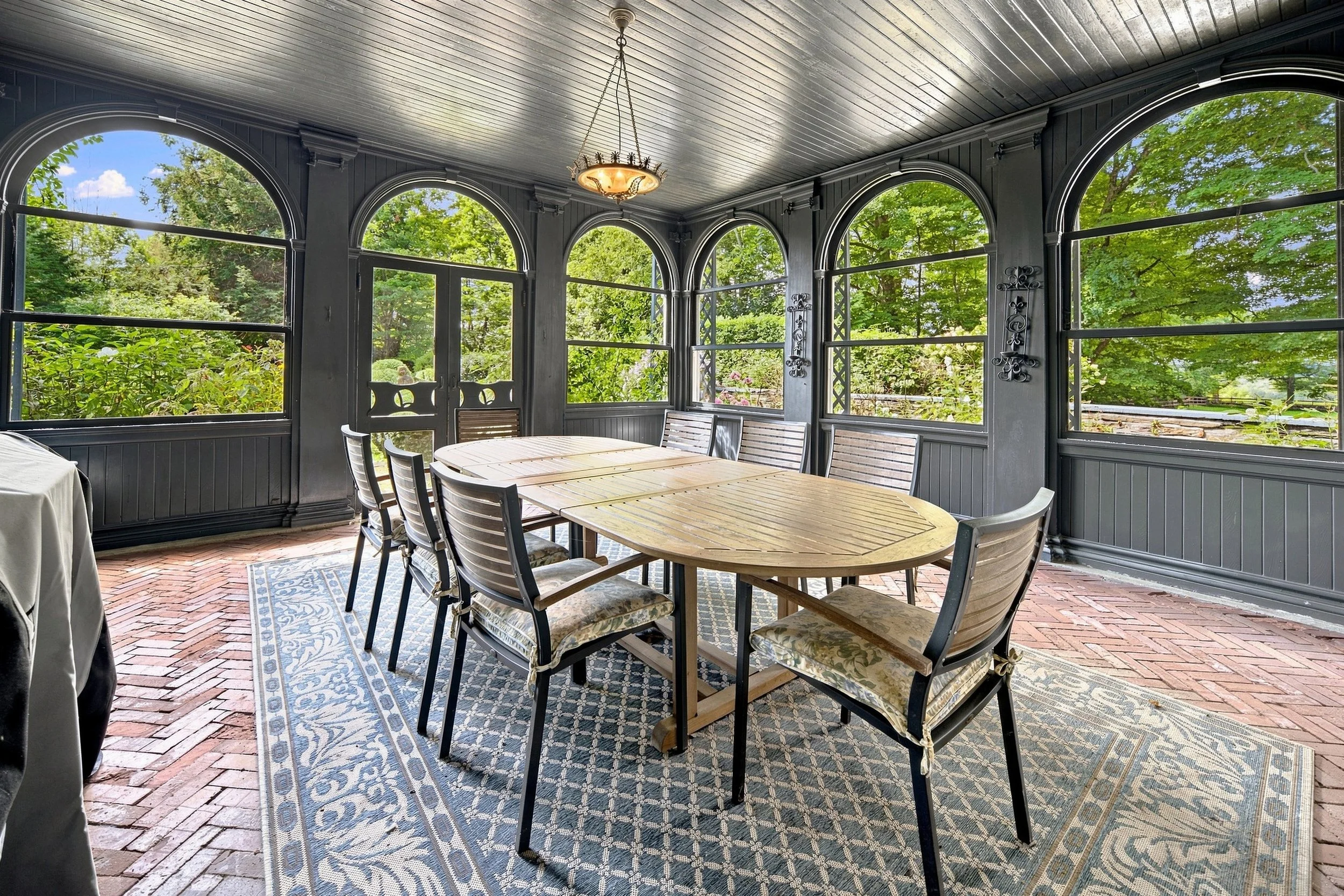 A screened porch with a wooden dining table and eight chairs, painted dark gray with decorative ironwork, overlooking a lush green garden with trees and plants.