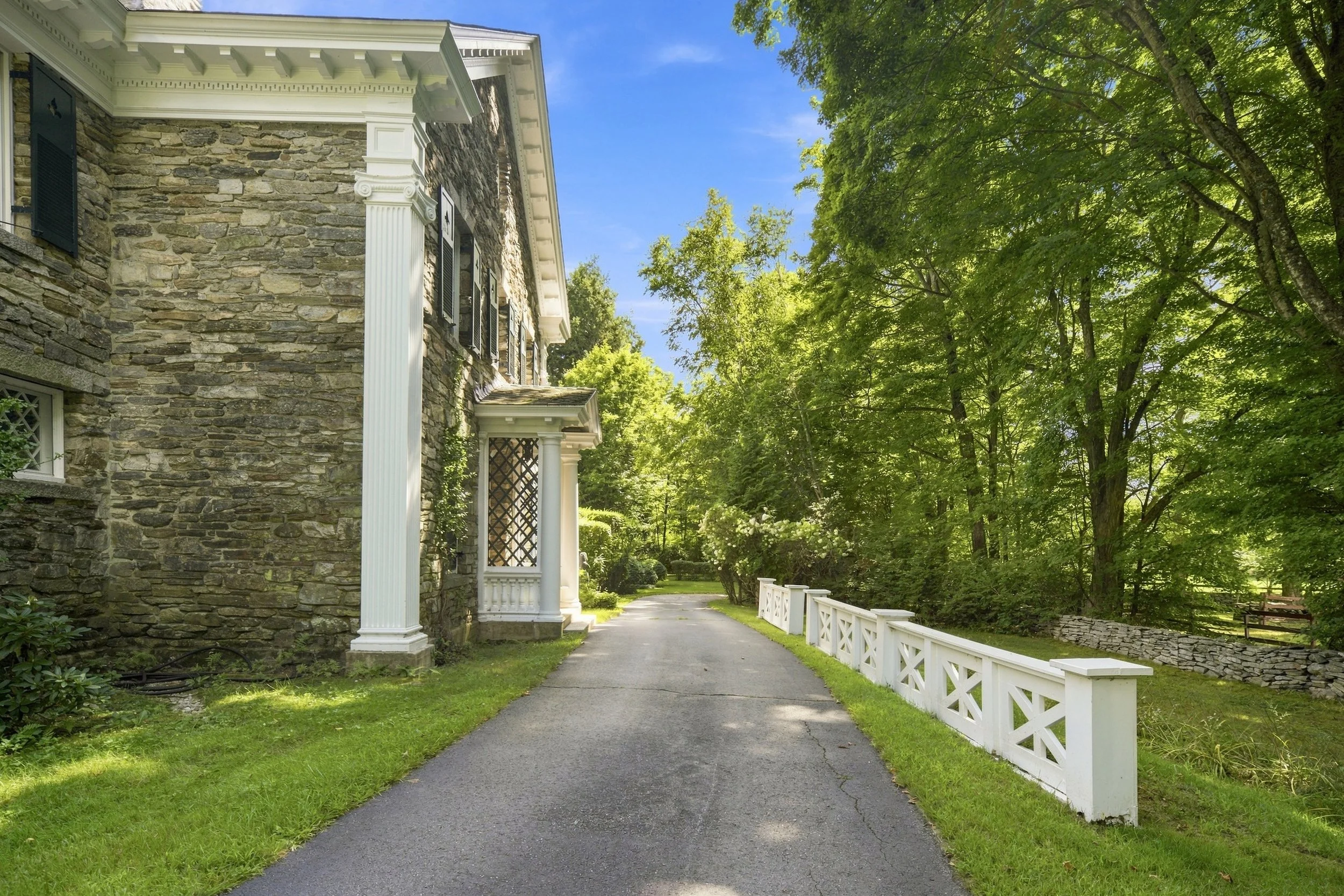 A stone house with black shutters and white trim, surrounded by lush green trees, with a paved walkway and white wooden fence leading into a landscaped yard.