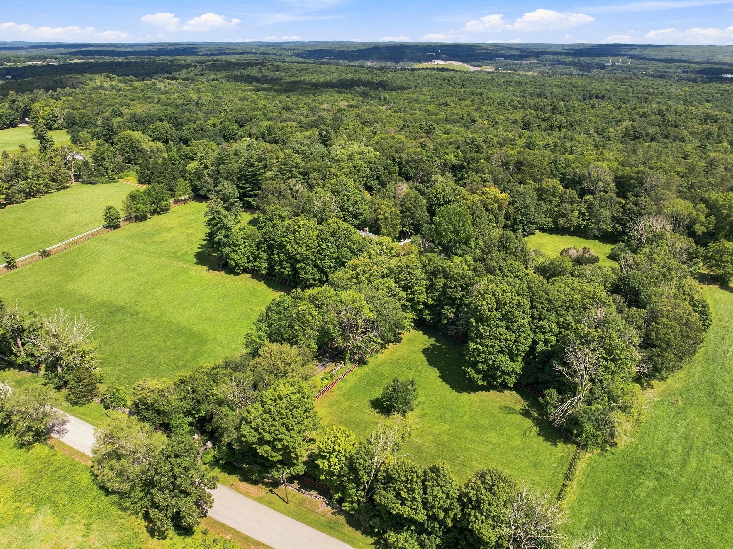Aerial view of a lush, green landscape with dense forested areas, open grassy fields, and a winding gravel path. The sky is blue with scattered clouds.