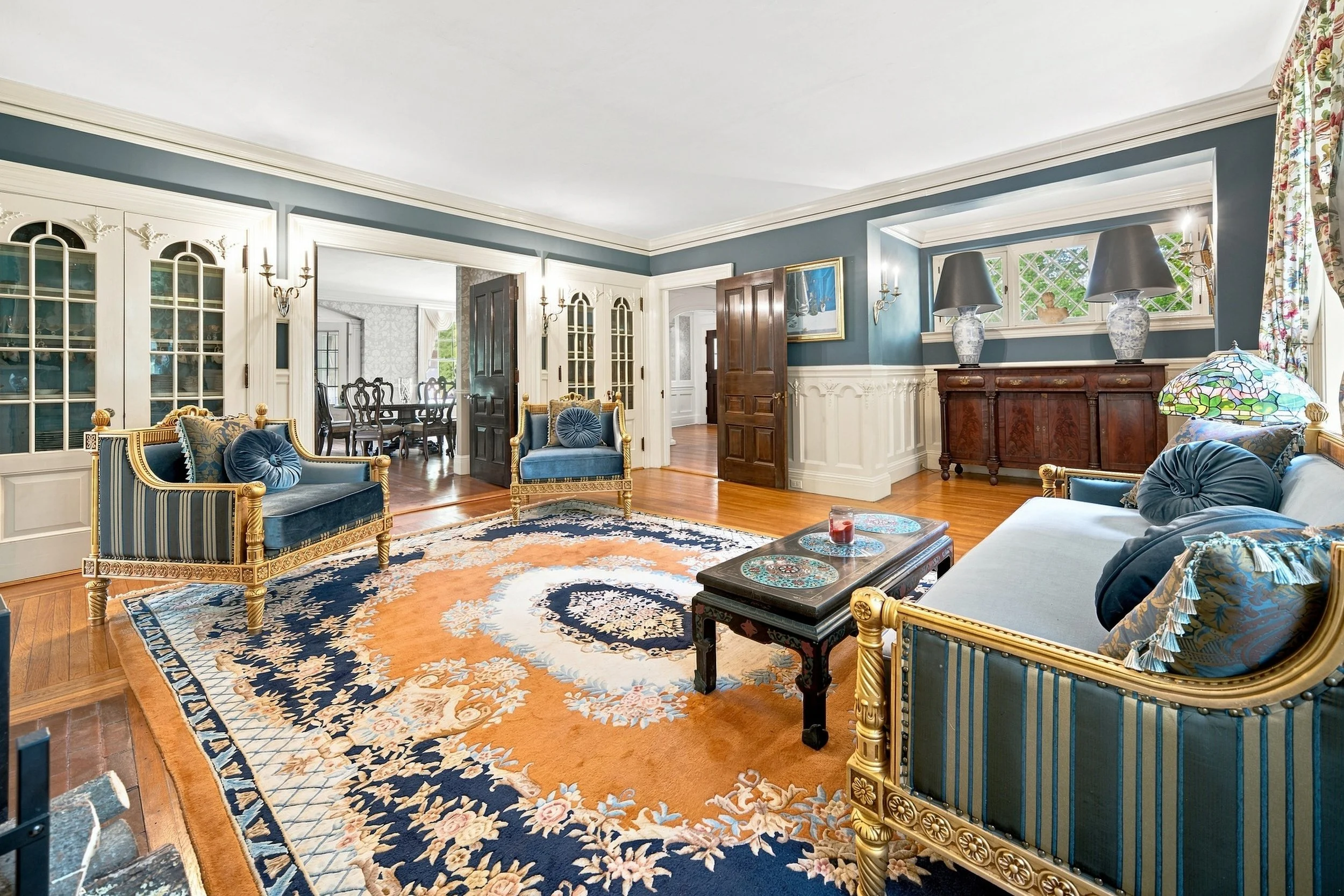 Living room with blue and gold furniture, ornate rugs, dark wood floors, and large windows with floral curtains.
