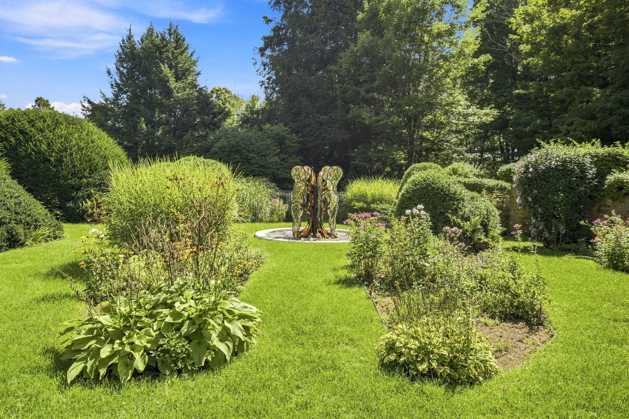 A lush green garden with various bushes and flowering plants surrounding a modern metal sculpture in the center, set under a bright blue sky with some clouds.
