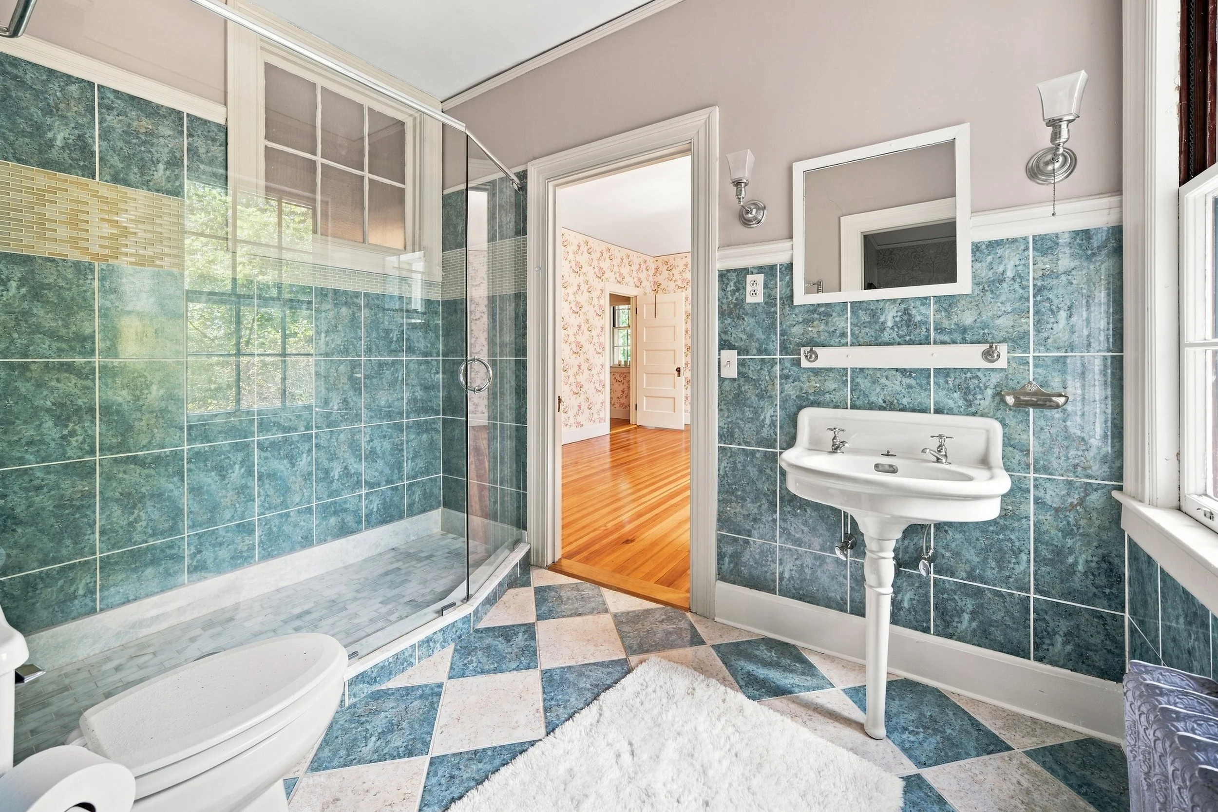 Bathroom with green and white tiled walls, a glass shower enclosure, a white sink with silver fixtures, and a mirror. There is a view into a room with floral wallpaper and wooden flooring.