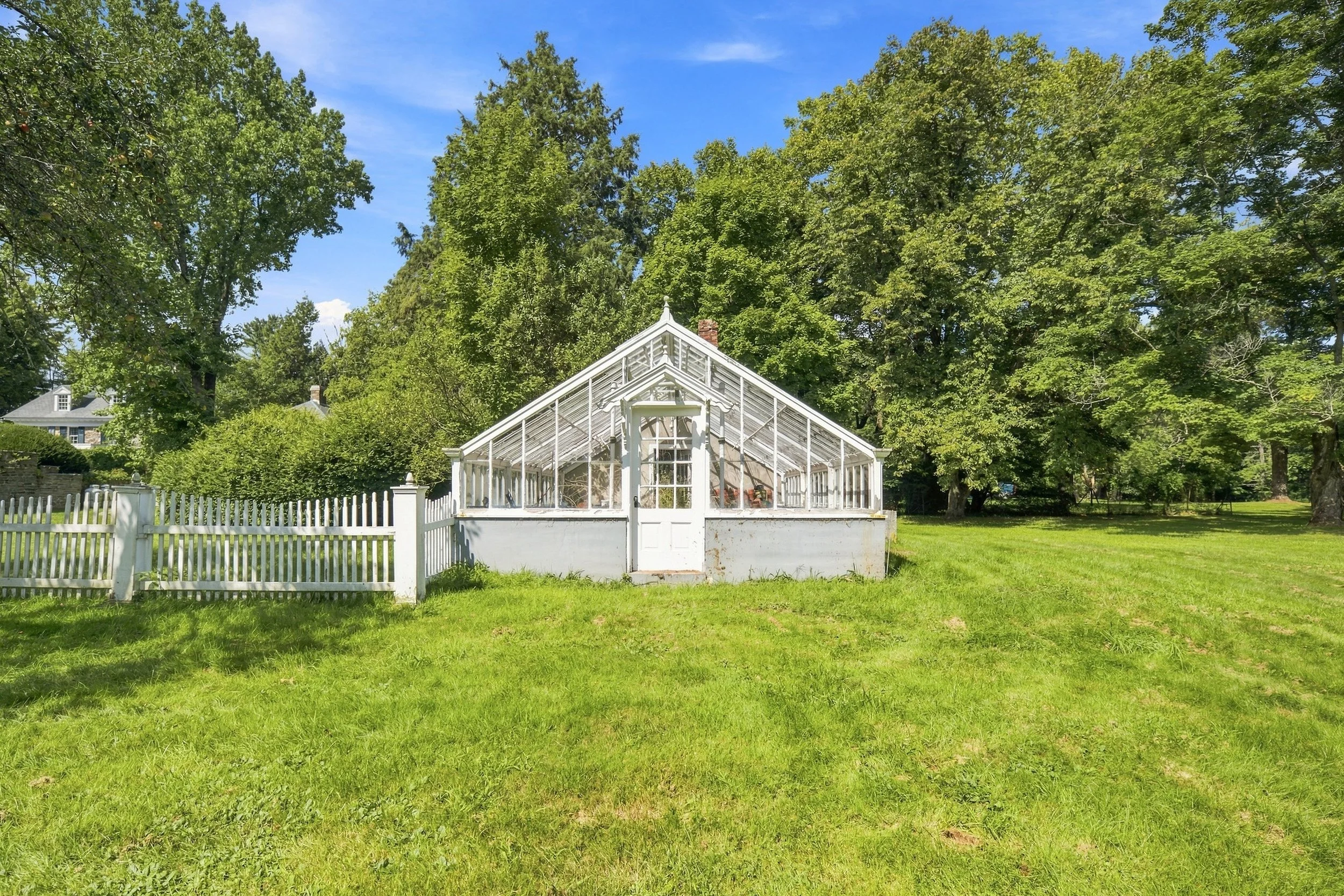 A white greenhouse with glass panels and a white picket fence in a grassy yard, surrounded by tall green trees under a blue sky.