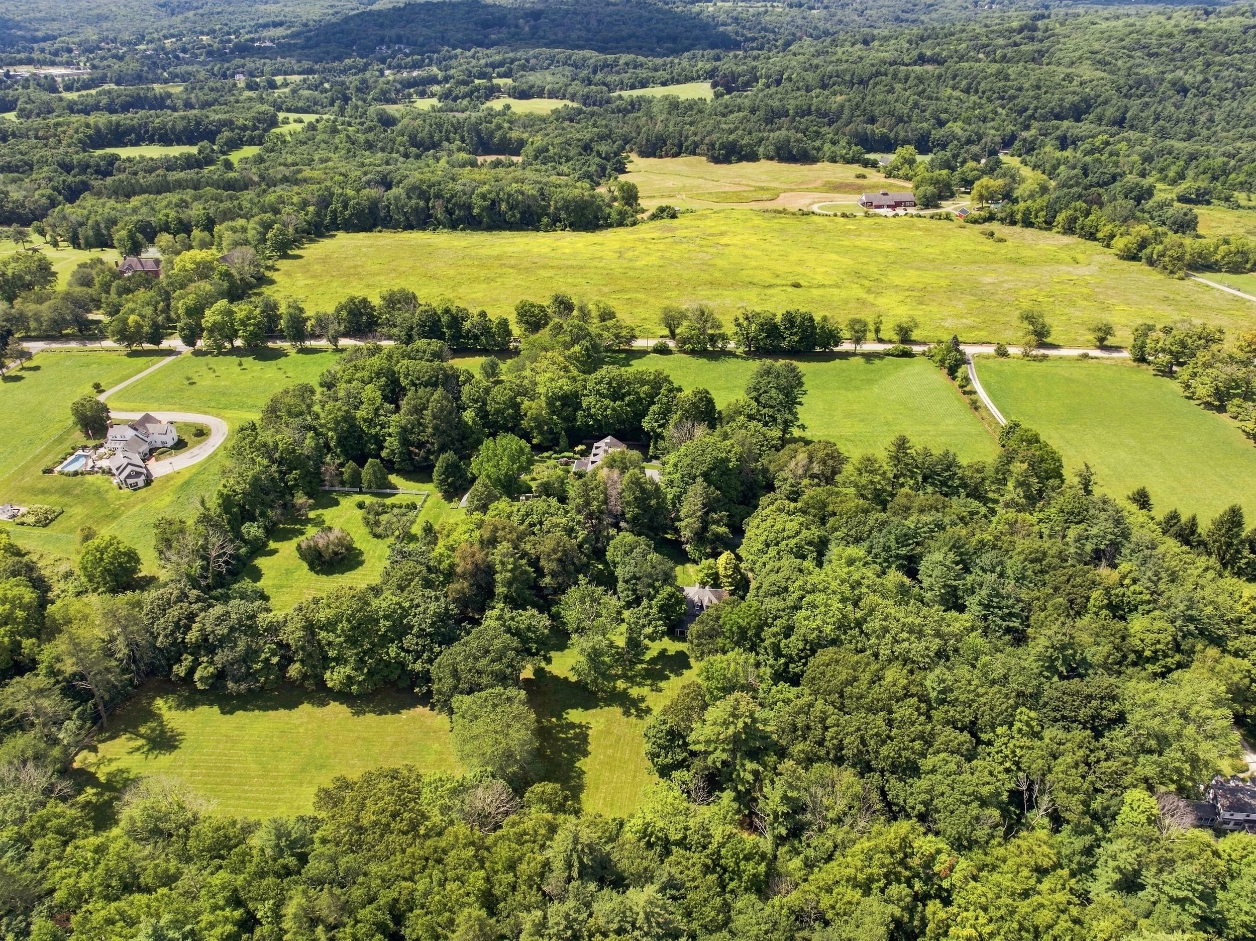 Aerial view of a rural landscape with green fields, trees, and scattered houses, including a house with a circular driveway and a swimming pool.