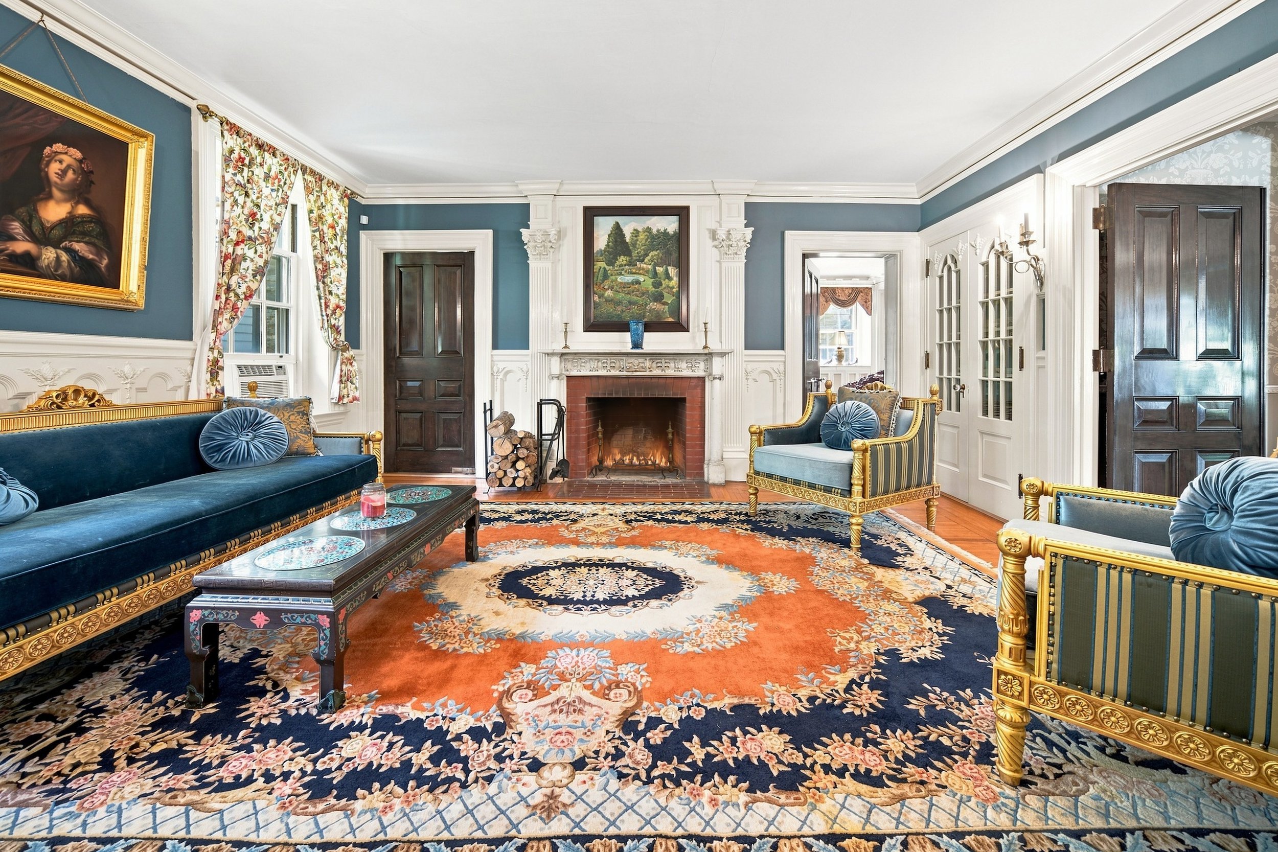 A room with a fireplace, floral curtain, ornate sofa, upholstered chairs, vibrant carpet, and classical artwork on the walls.