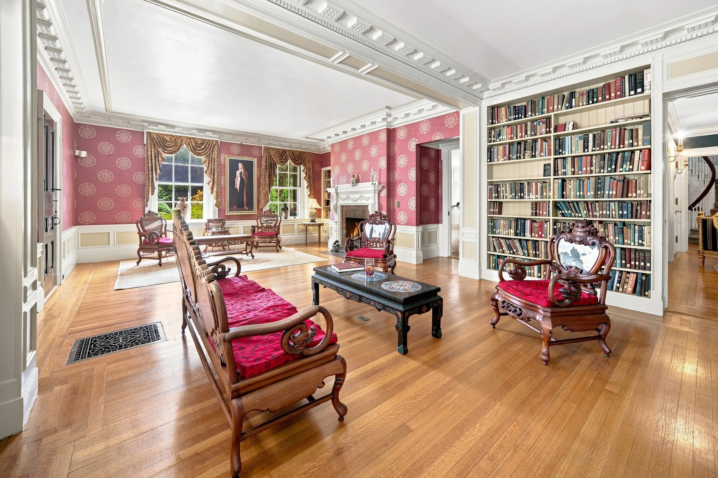 A spacious living room with pink wallpaper featuring circular patterns, white crown molding, and hardwood floors. It contains a white fireplace with a portrait hanging above it, several chairs with red upholstery, a large bookshelf filled with books,