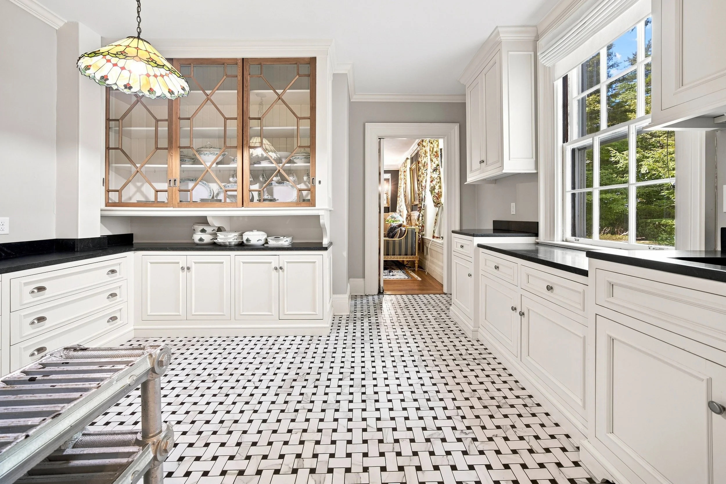 A kitchen with white cabinets, black countertops, a black-and-white patterned tile floor, a large window showing greenery outside, and a wooden glass-paneled cabinet in the background.