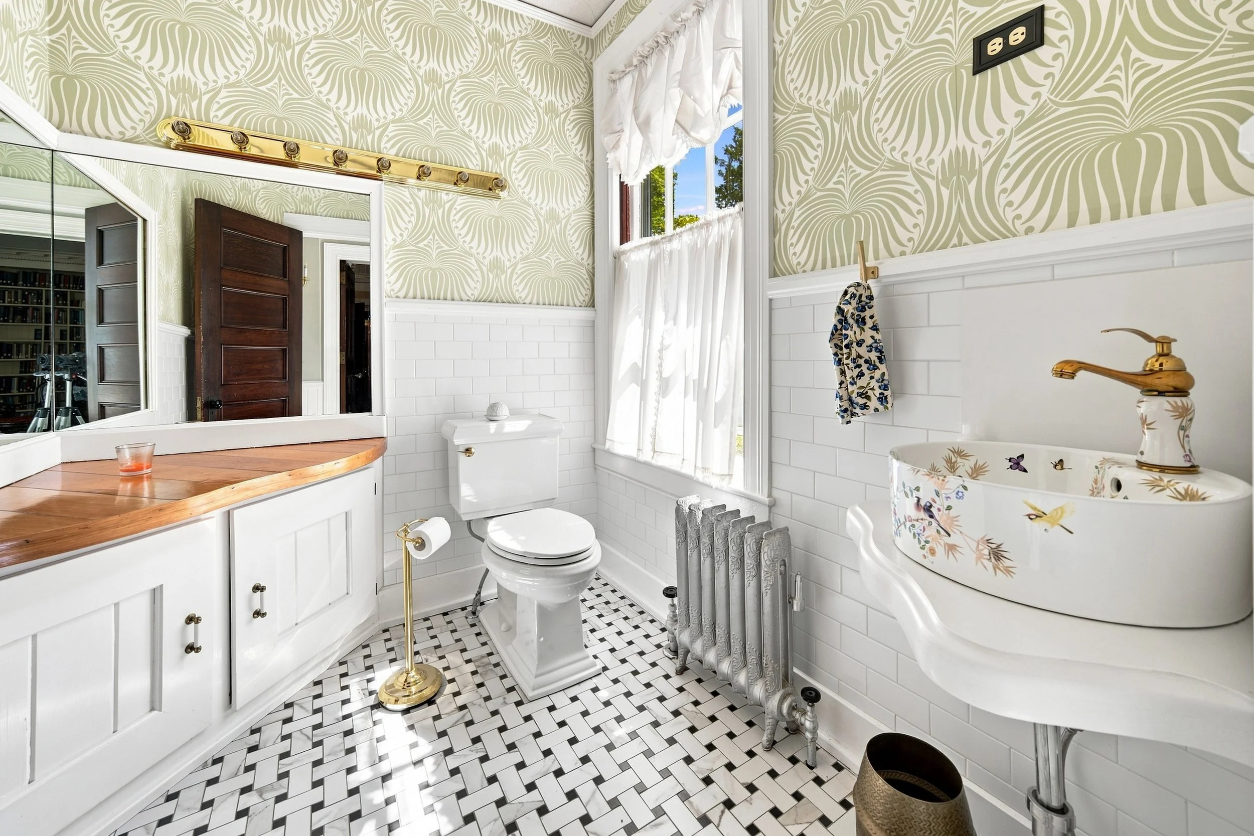 A vintage-inspired bathroom with patterned wallpaper, a window with white curtains, a white toilet, a white tiled sink with a floral design, a towel hanging on a hook, and a small radiator with a patterned towel. The floor has a black and white geome