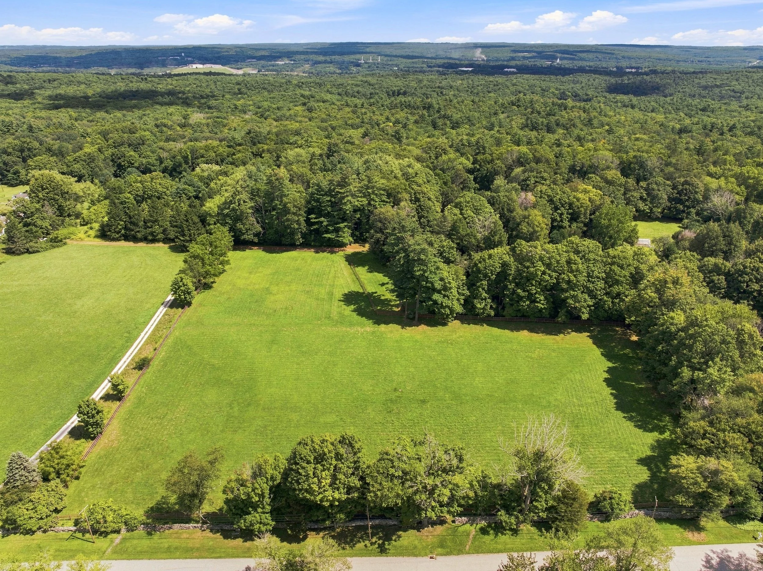 An aerial view of a lush green landscape with dense forest, open fields, and distant hills under a partly cloudy sky.