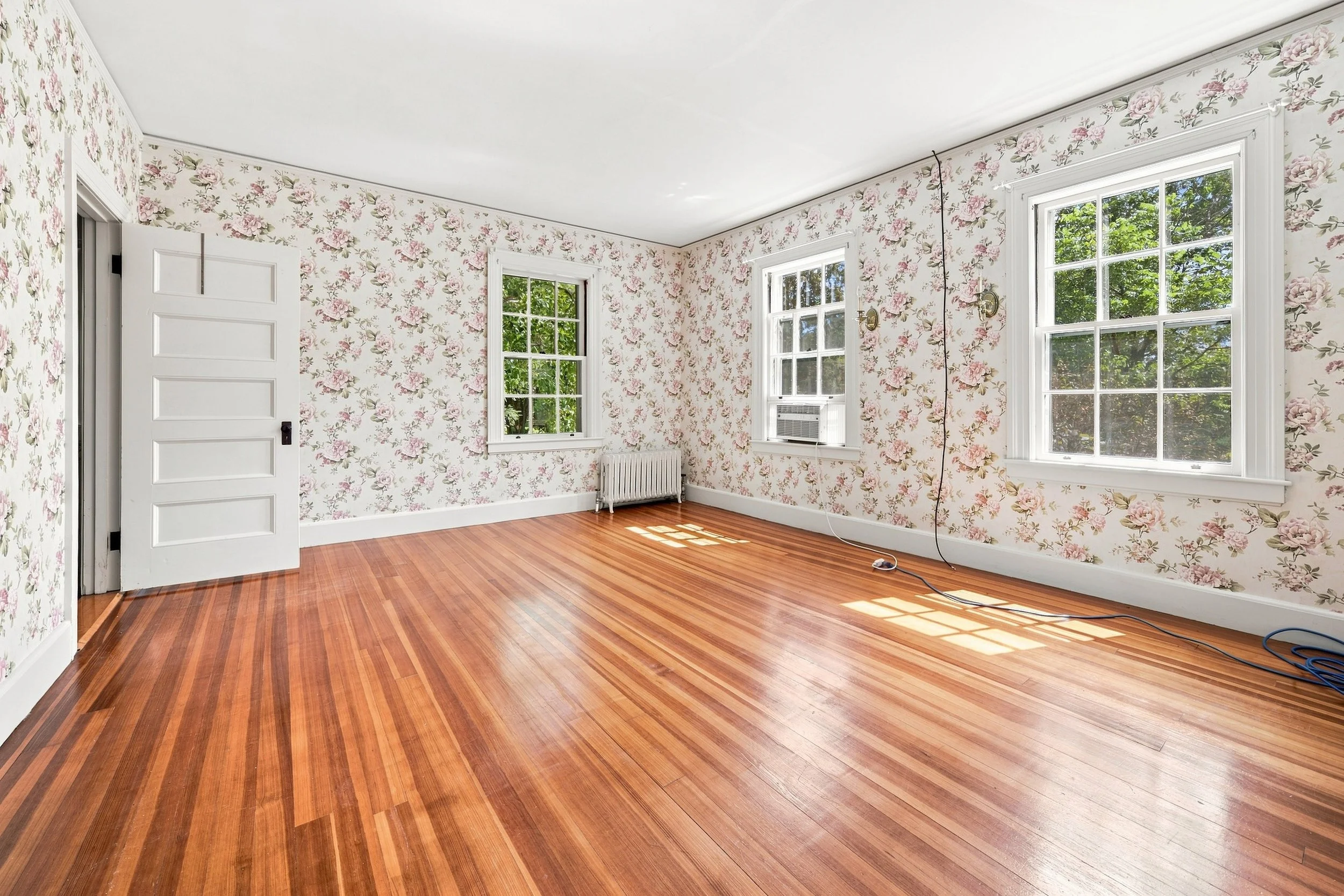 Empty room with wooden floors, floral wallpaper, three large windows, and an open white door.