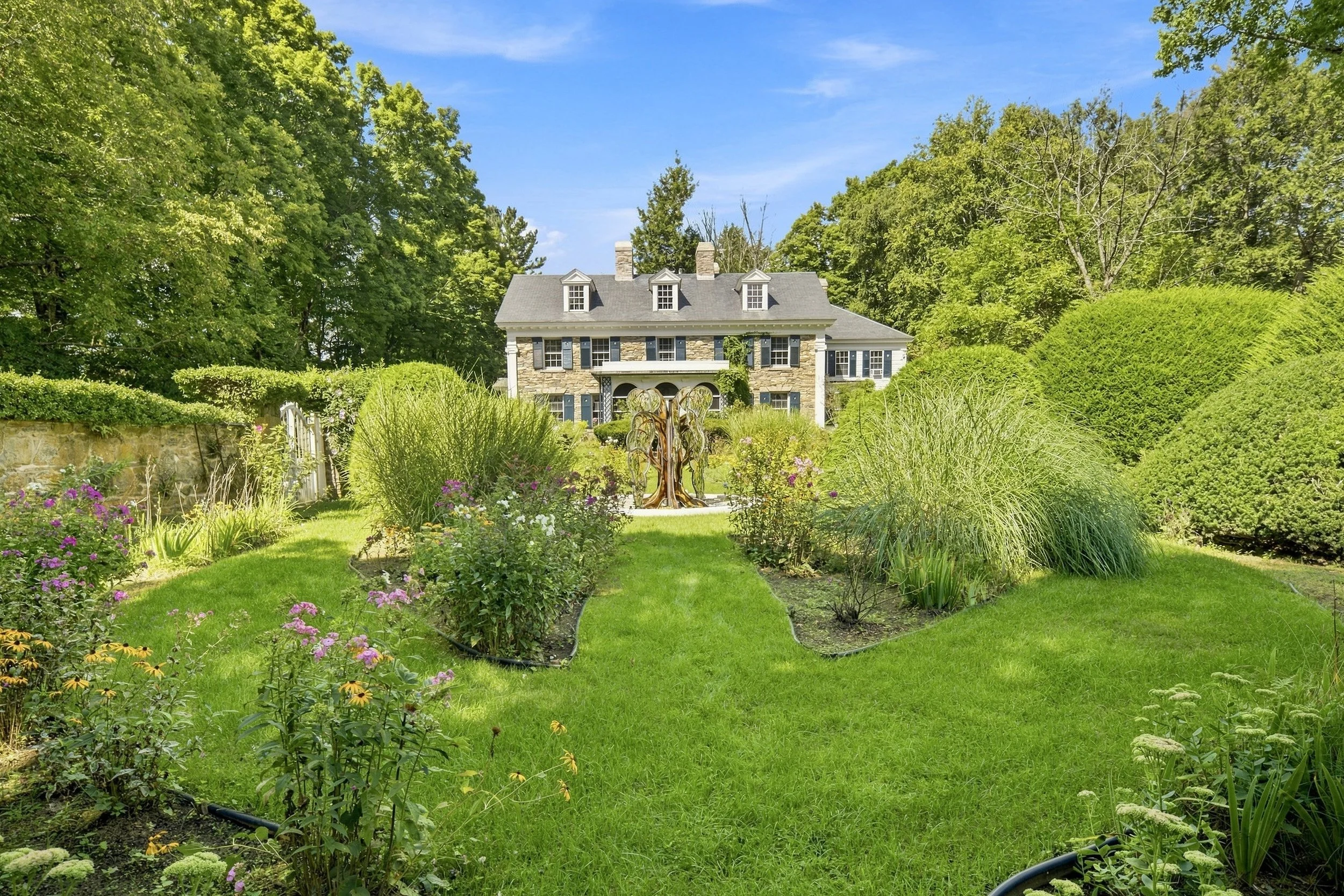 Front view of a large, historic house surrounded by a well-maintained garden with lush green trees and flowering plants.
