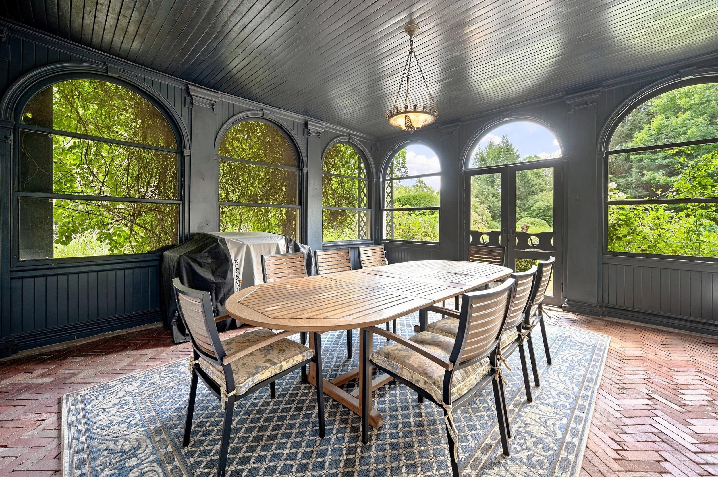 Enclosed porch with a wooden dining table and chairs, surrounded by large windows showing lush greenery outside, decorated with a vintage chandelier and patterned rug.