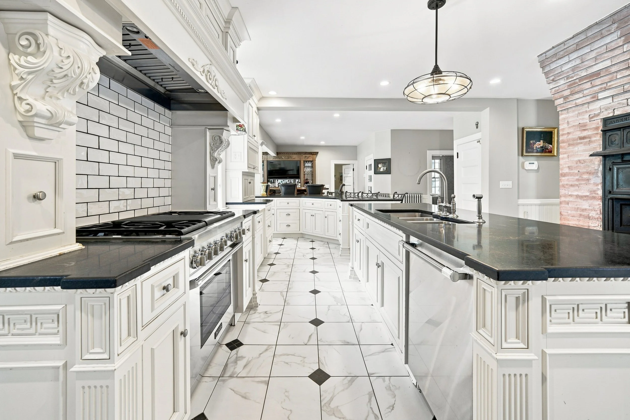 White kitchen with black countertops, white cabinetry with decorative molding, stainless steel appliances, a brick wall, and a pendant ceiling light.