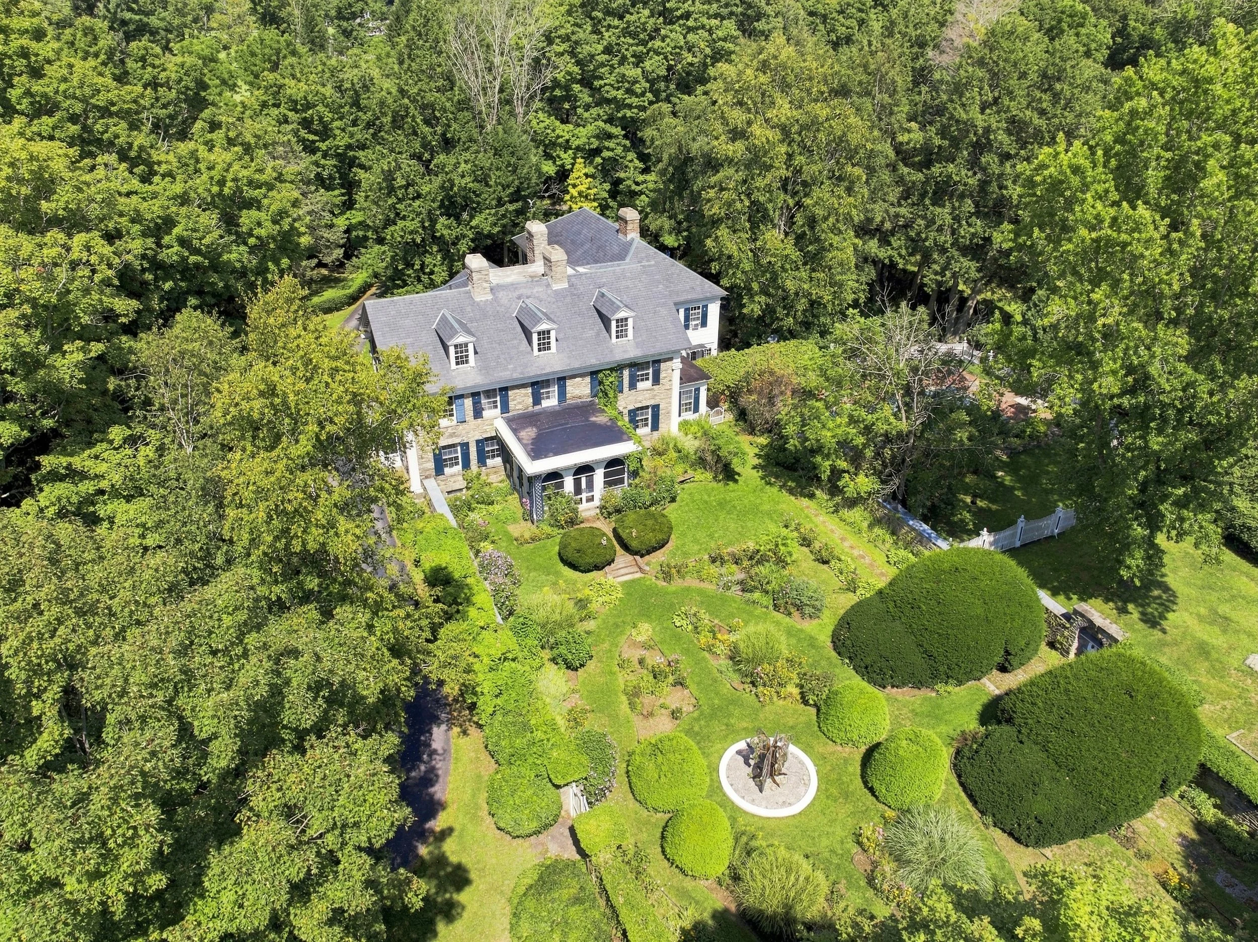 An aerial view of a large house surrounded by lush green trees and a well-maintained garden with trimmed bushes and a statue in the front yard.