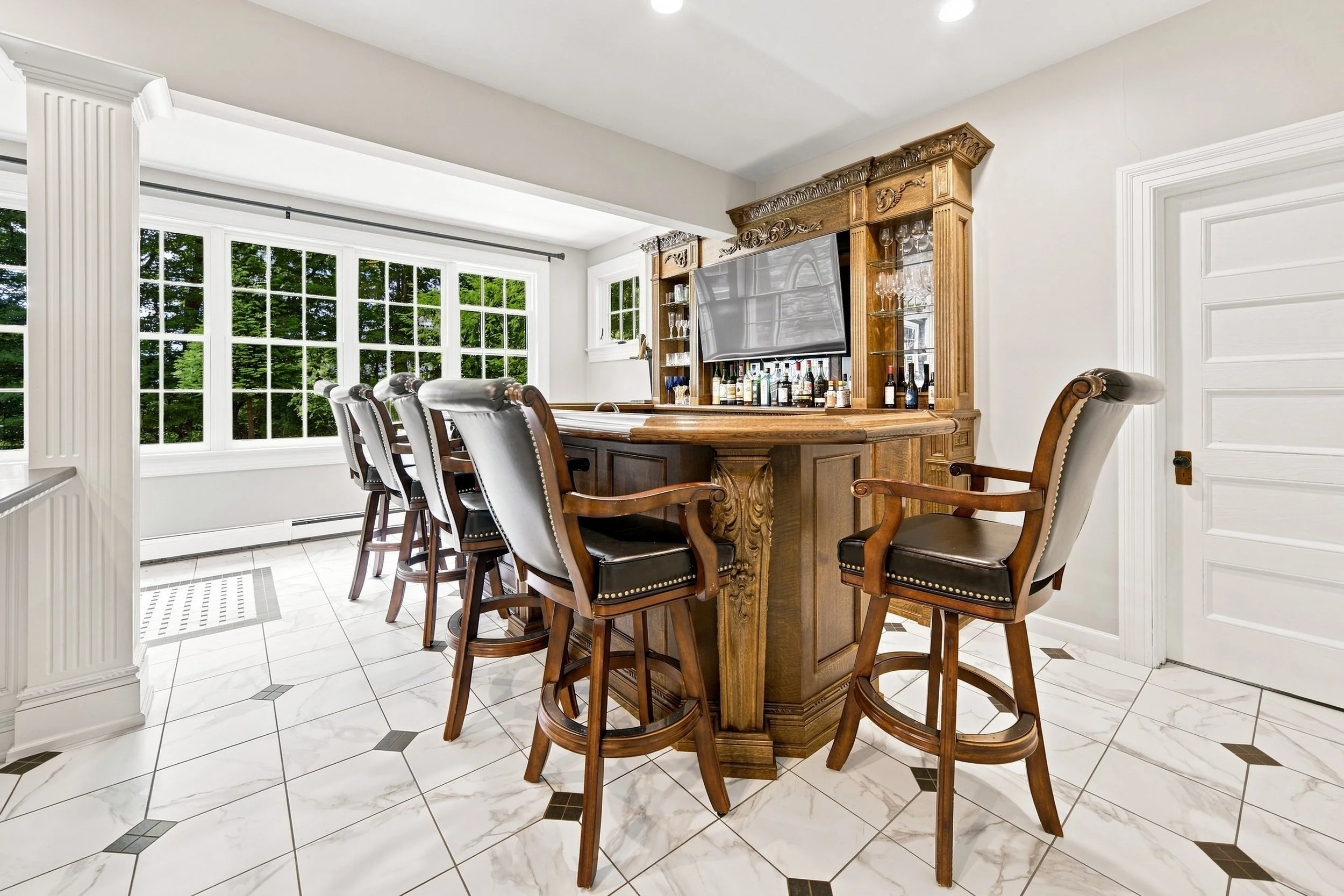 A room with a large window, a wooden bar table with chairs, and a wooden cabinet with glassware and bottles, featuring a TV mounted on it.