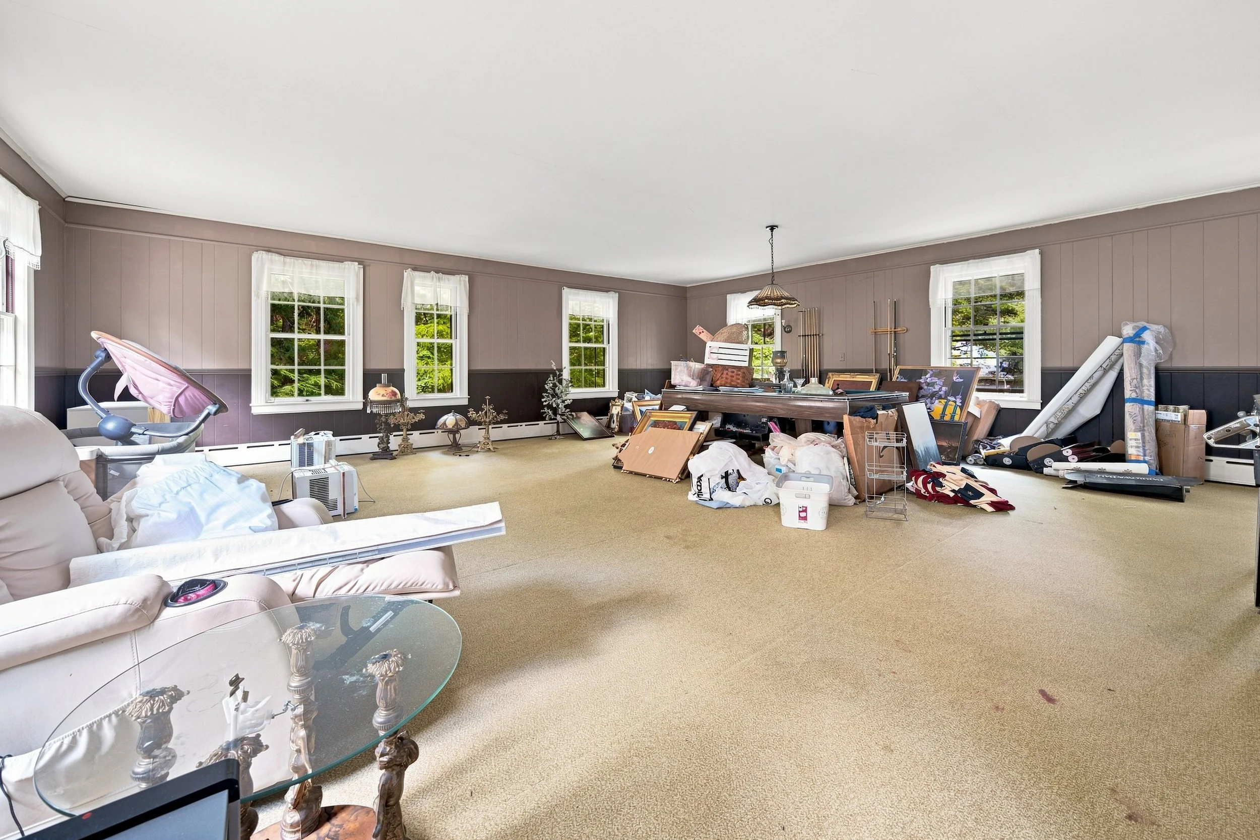 Living room cluttered with various items, including furniture, picture frames, and boxes, with three windows providing natural light.