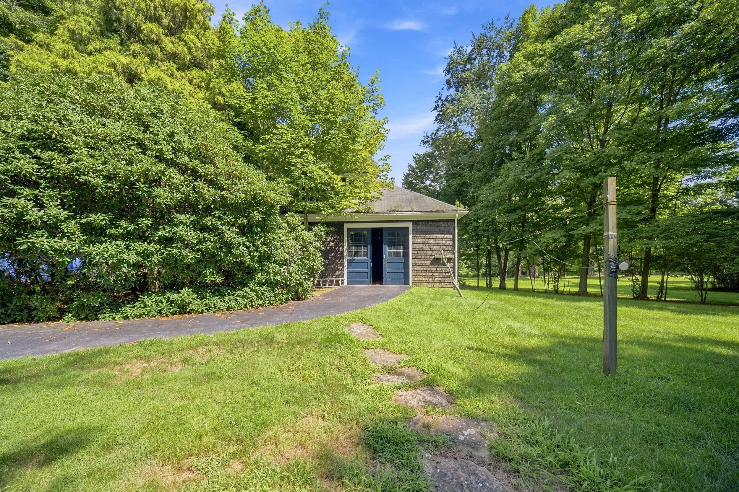 A suburban backyard with a small shed that has gray shingles and blue double doors, surrounded by green trees, a grassy lawn, and a blue sky with some clouds. There is a utility pole with wires on the right side of the image.