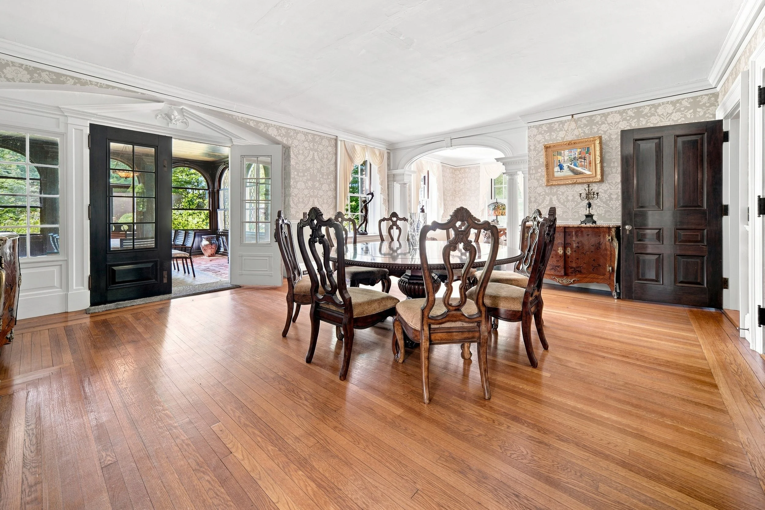 A dining room with a wooden oval table and six ornate wooden chairs, dark doors, large windows, and light-colored wallpaper with floral patterns.