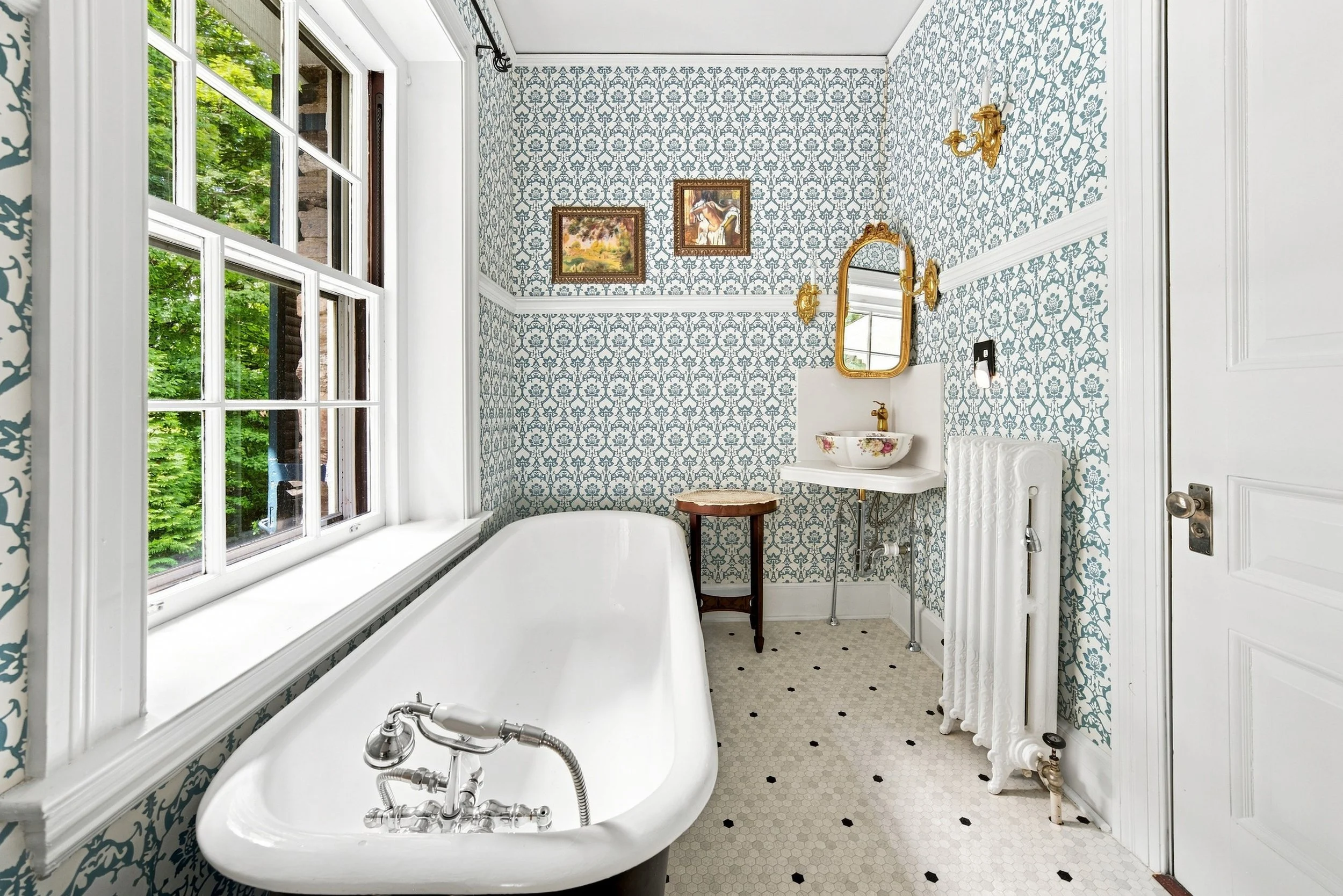 Vintage bathroom with a bathtub near a large window, blue floral wallpaper, a small sink with a mirror, and decorative wall sconces.