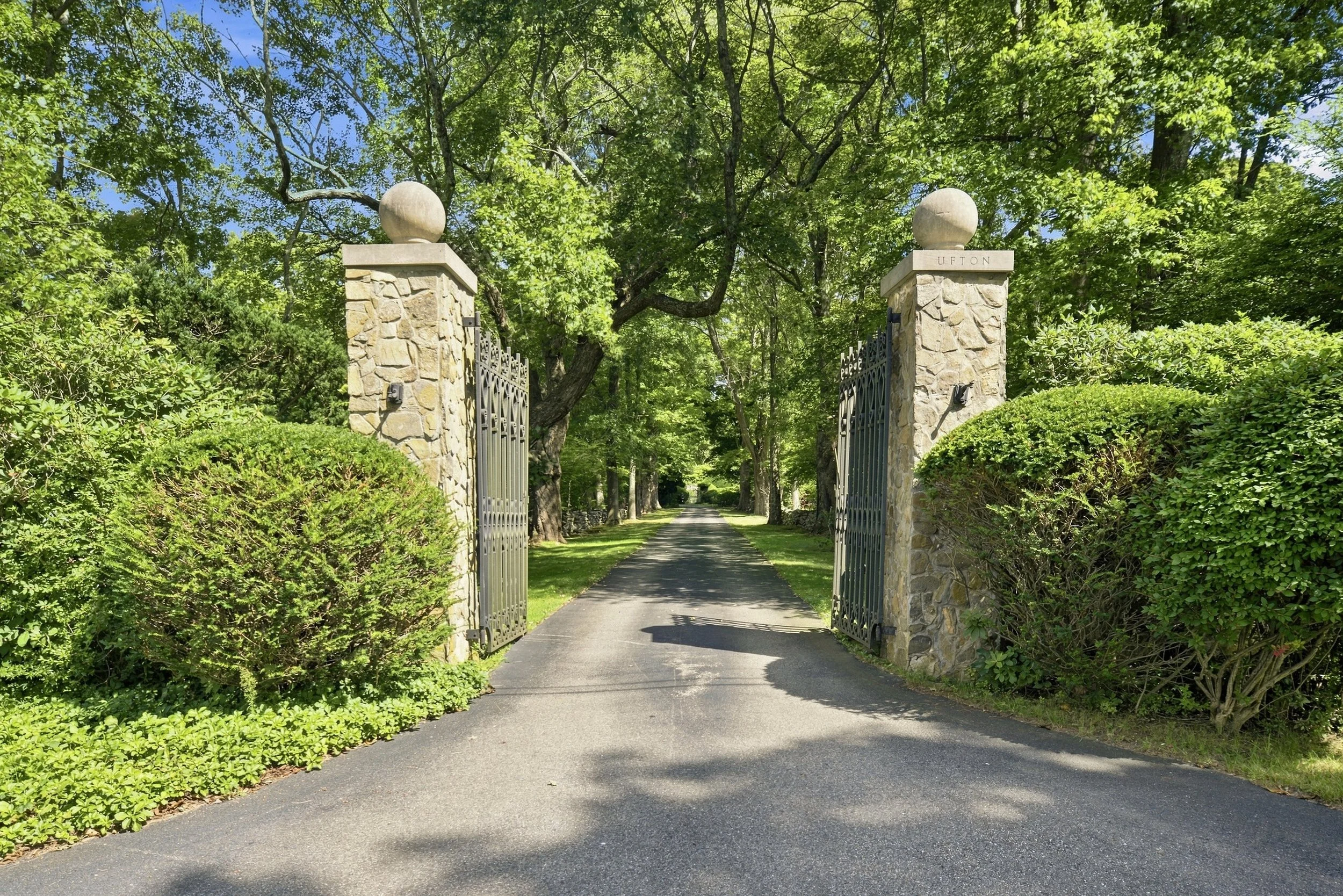 The image shows the entrance to a lush, green park with stone pillars and metal gates. A driveway extends through the gates into a tree-lined pathway. The stone pillar on the right has a sign with the name "Upton". The area is surrounded by dense tre