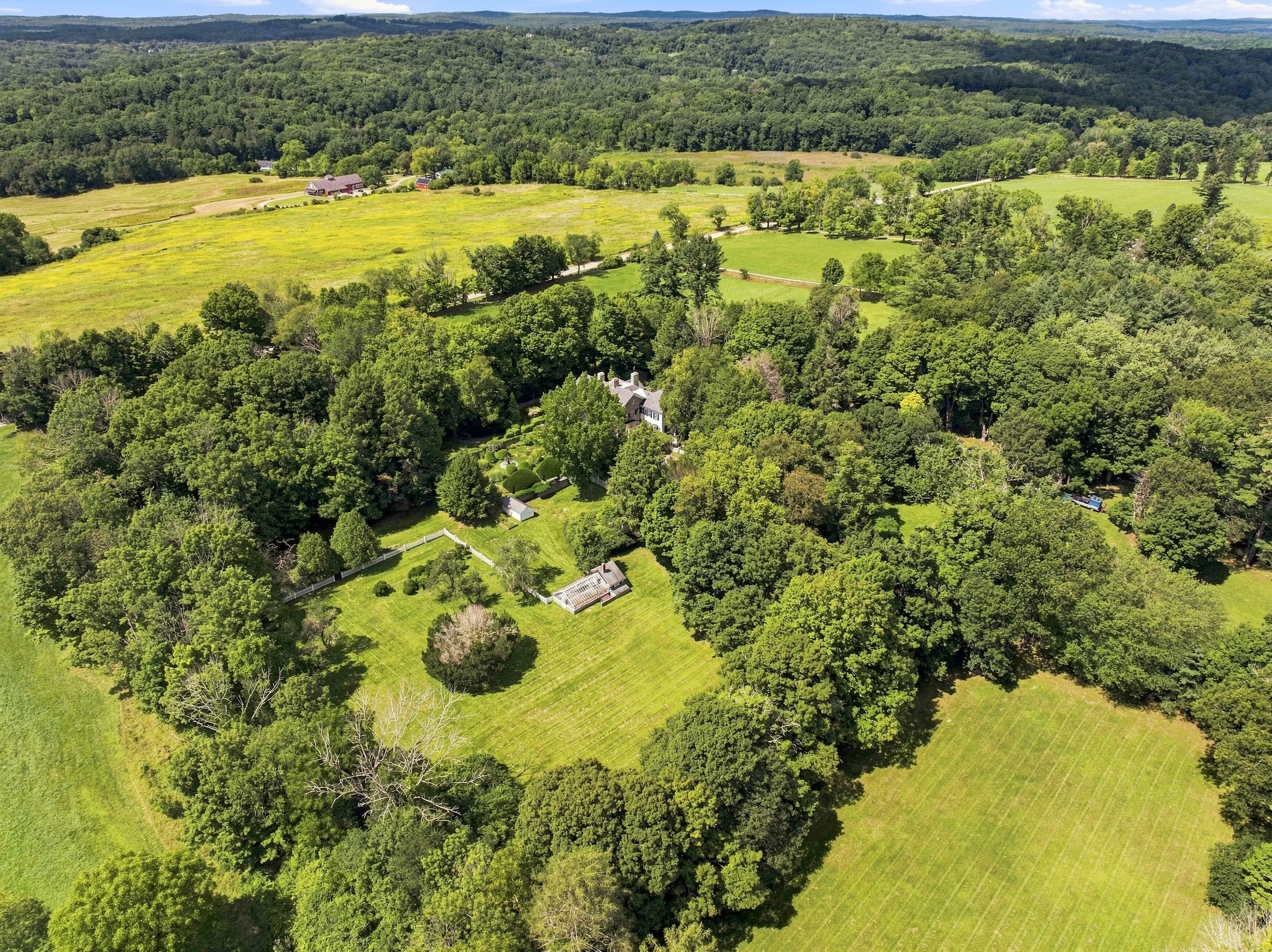 Aerial view of lush green fields, trees, and distant hills, with several houses and a farm in a rural landscape.