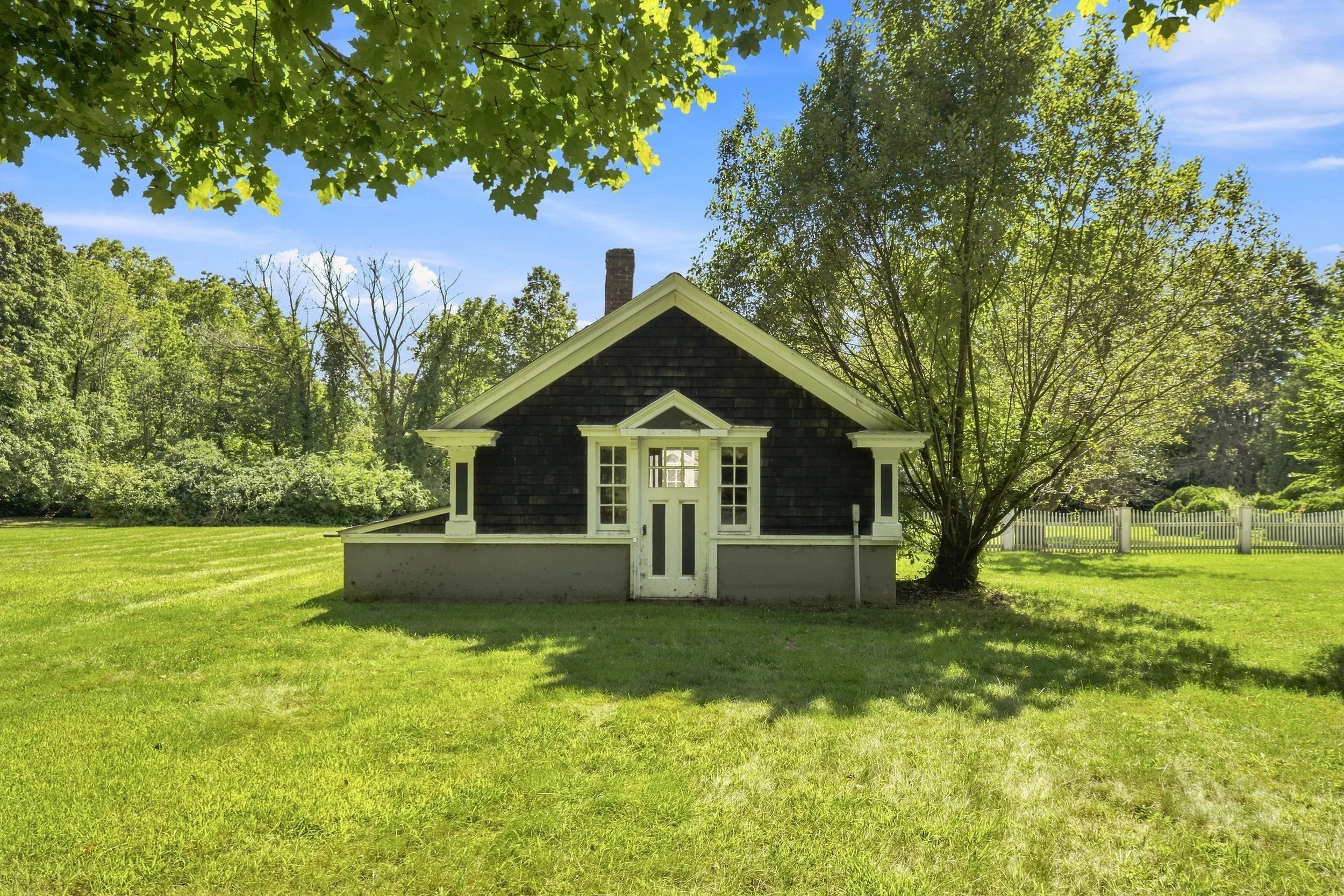 A small house with dark shingles on the roof and white trim, surrounded by green trees and a well-maintained lawn under a bright blue sky.