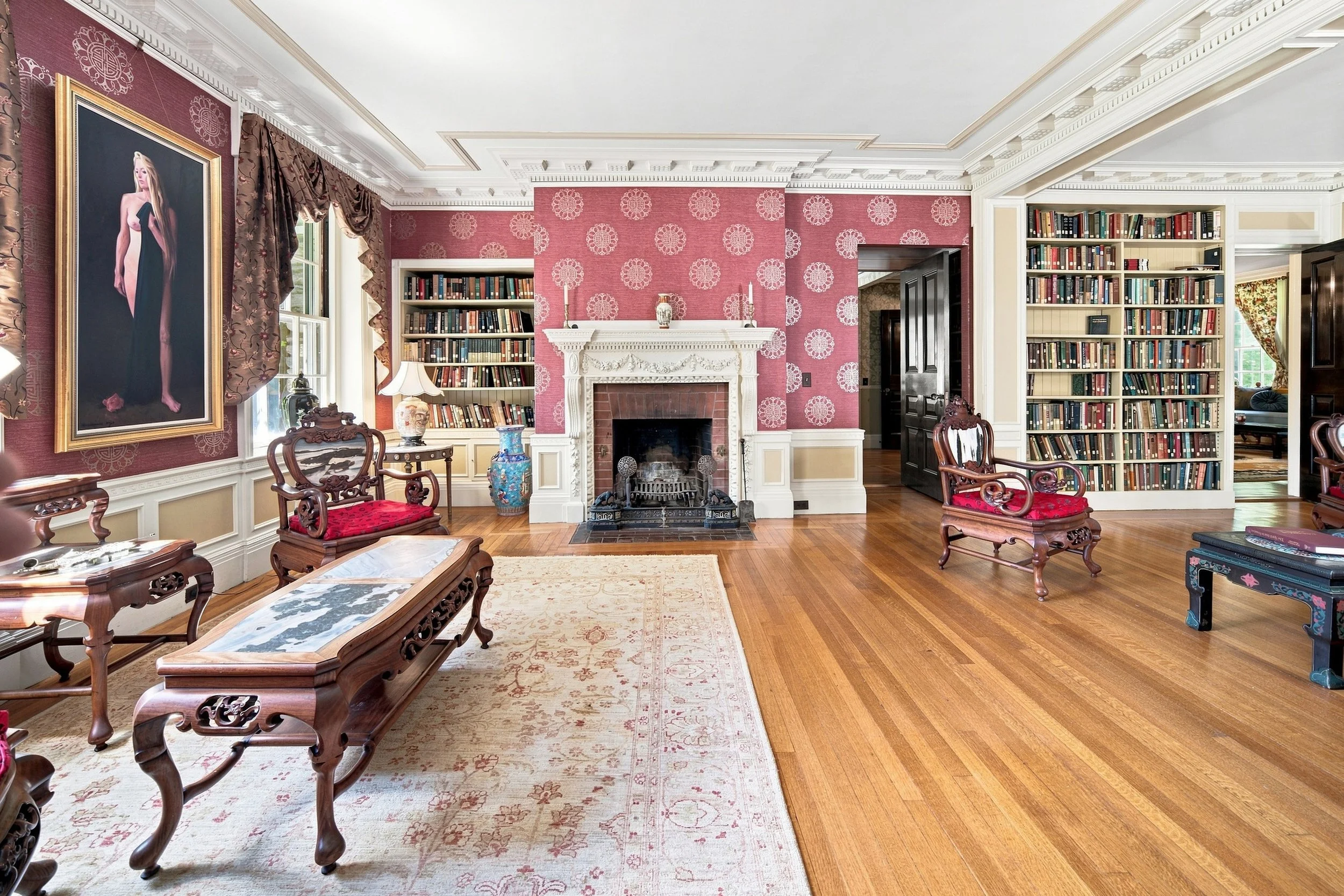 A living room with red wallpaper, white crown molding, and a fireplace. There are bookshelves on either side of the fireplace, and a portrait of a woman hanging on the wall. The room has wooden floors, a beige area rug, and vintage wooden chairs with