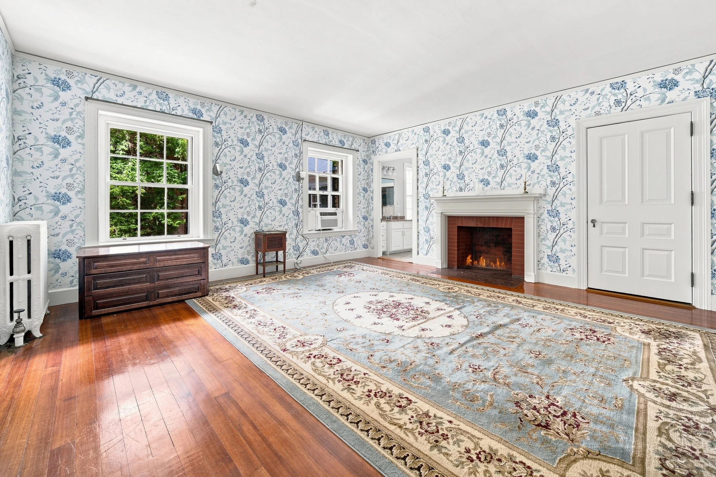 Living room with floral wallpaper, hardwood floor, a large area rug, a fireplace, and windows showing green trees outside.