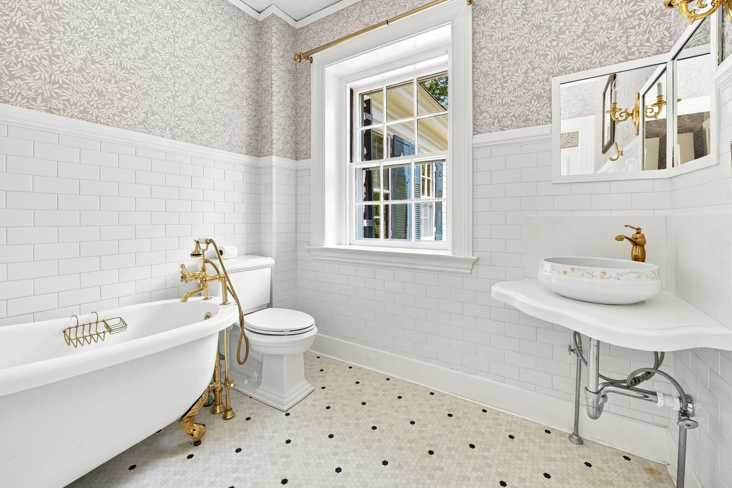 A white clawfoot bathtub with gold fixtures next to a toilet in a bathroom with white subway tile on the walls, a window with shutters, and a mirror above a white sink with a gold faucet.