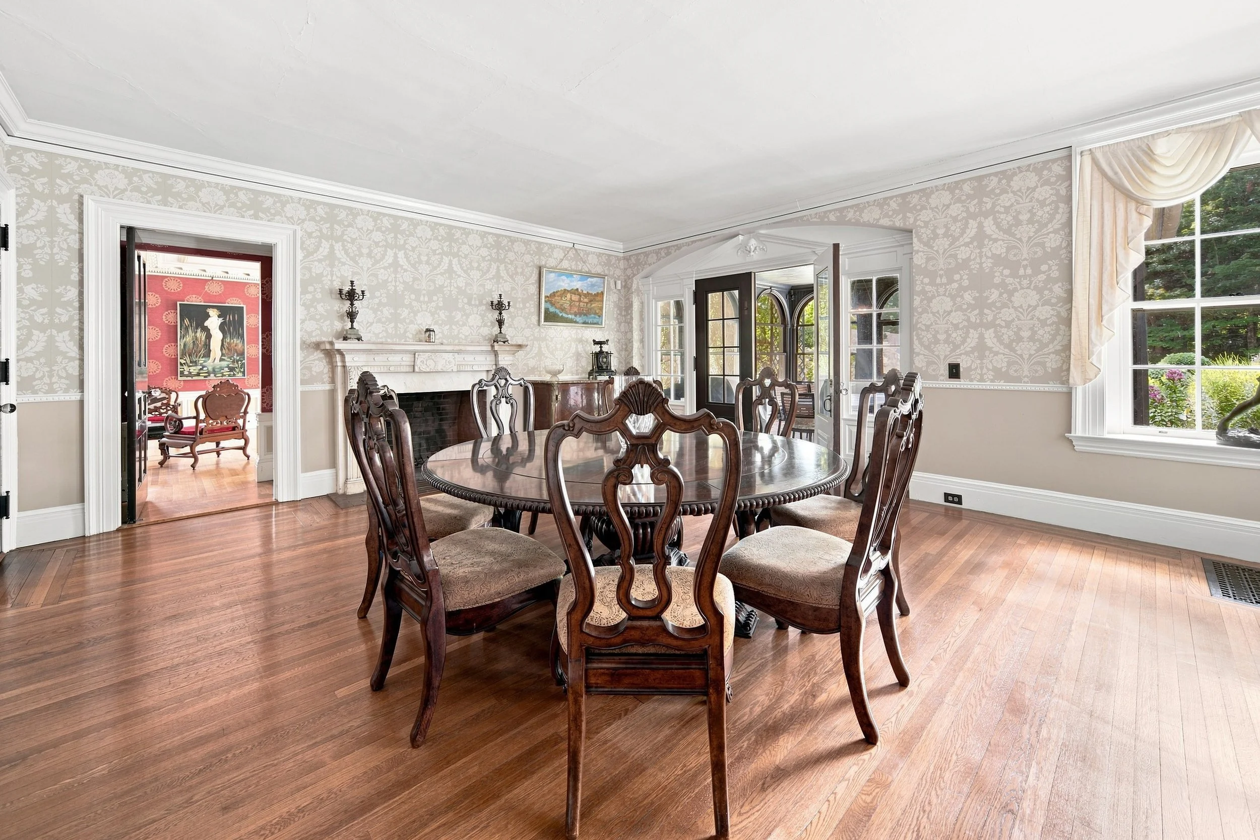 A dining room with a round wooden table and eight matching chairs with upholstered seats. The walls are decorated with patterned wallpaper, and large windows with curtains let in natural light. There is a fireplace on one wall with artwork above it, 