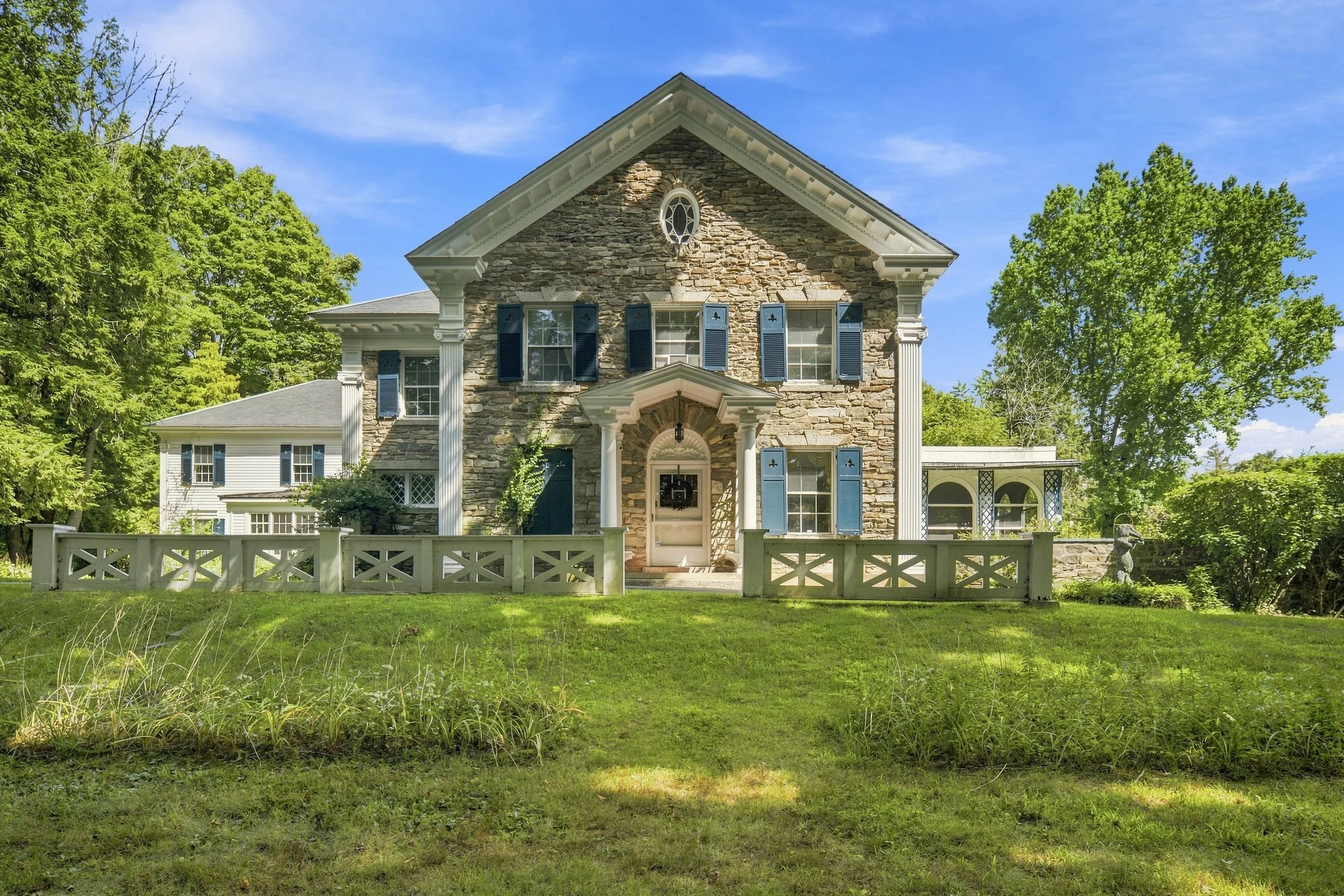 Front view of a large house with a stone facade, white columns, and blue shutters, surrounded by green trees and a white fence, under a blue sky.