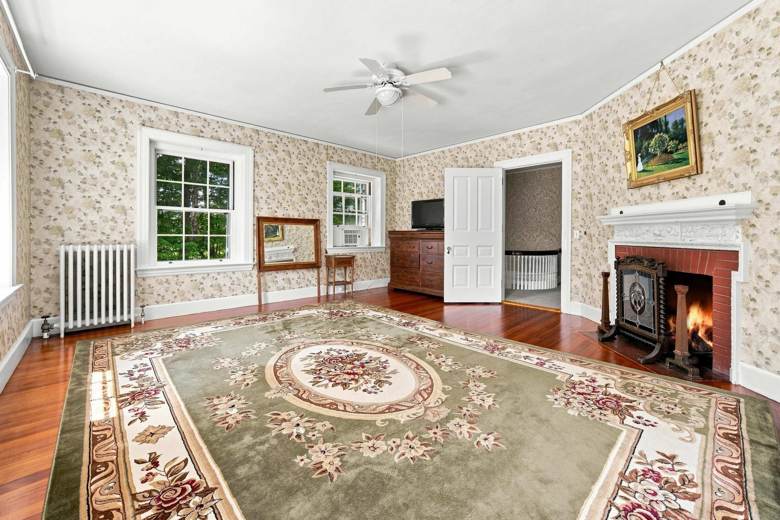Living room with floral wallpaper, hardwood floor, a fireplace with a white mantel, a large area rug with floral designs, a ceiling fan, and a window showing green trees outside.