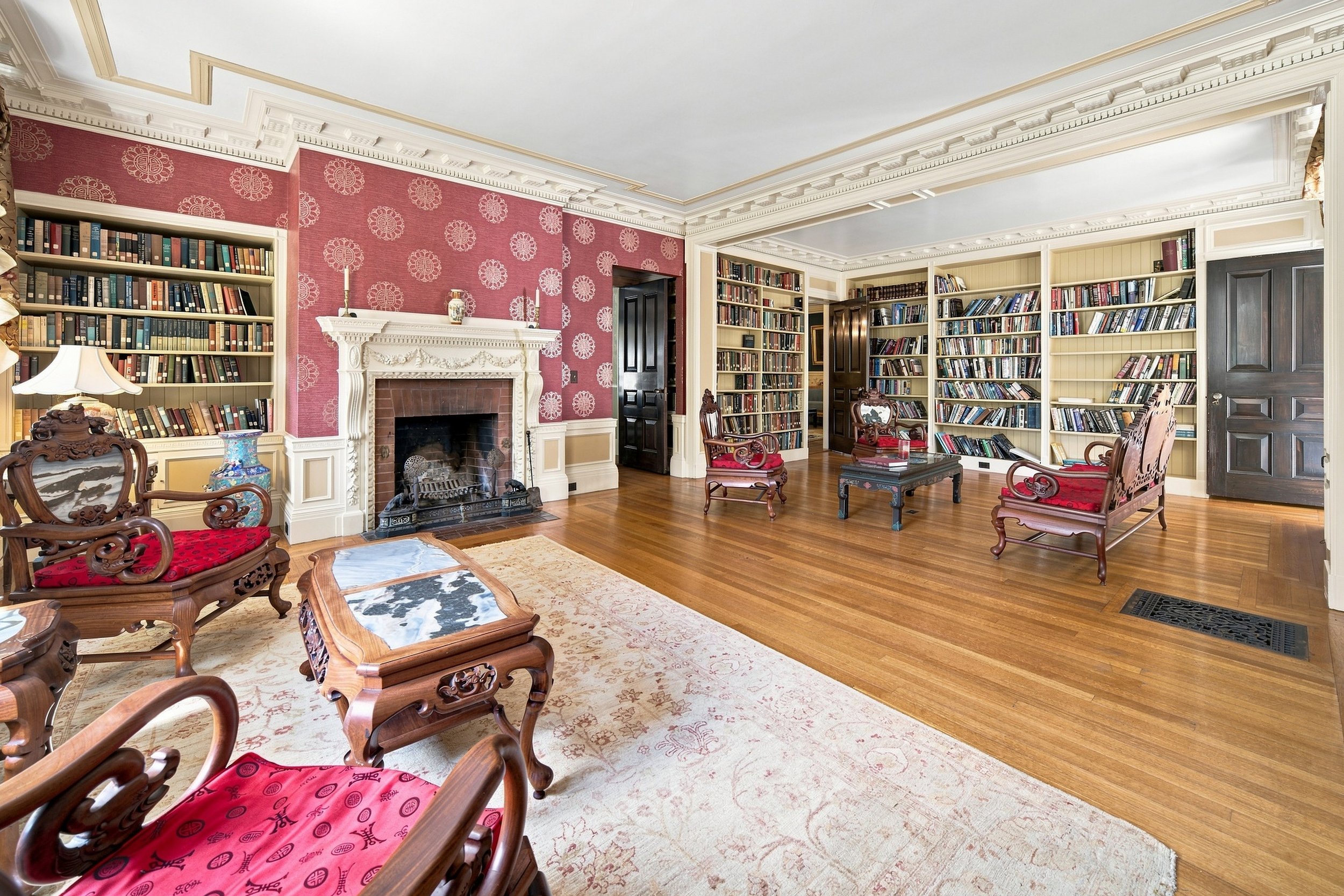 A spacious traditional-style living room with built-in bookshelves filled with books, a decorative fireplace, ornate wooden chairs with red padded seats, and an area rug on hardwood flooring. The room features crown molding and a ceiling with intrica
