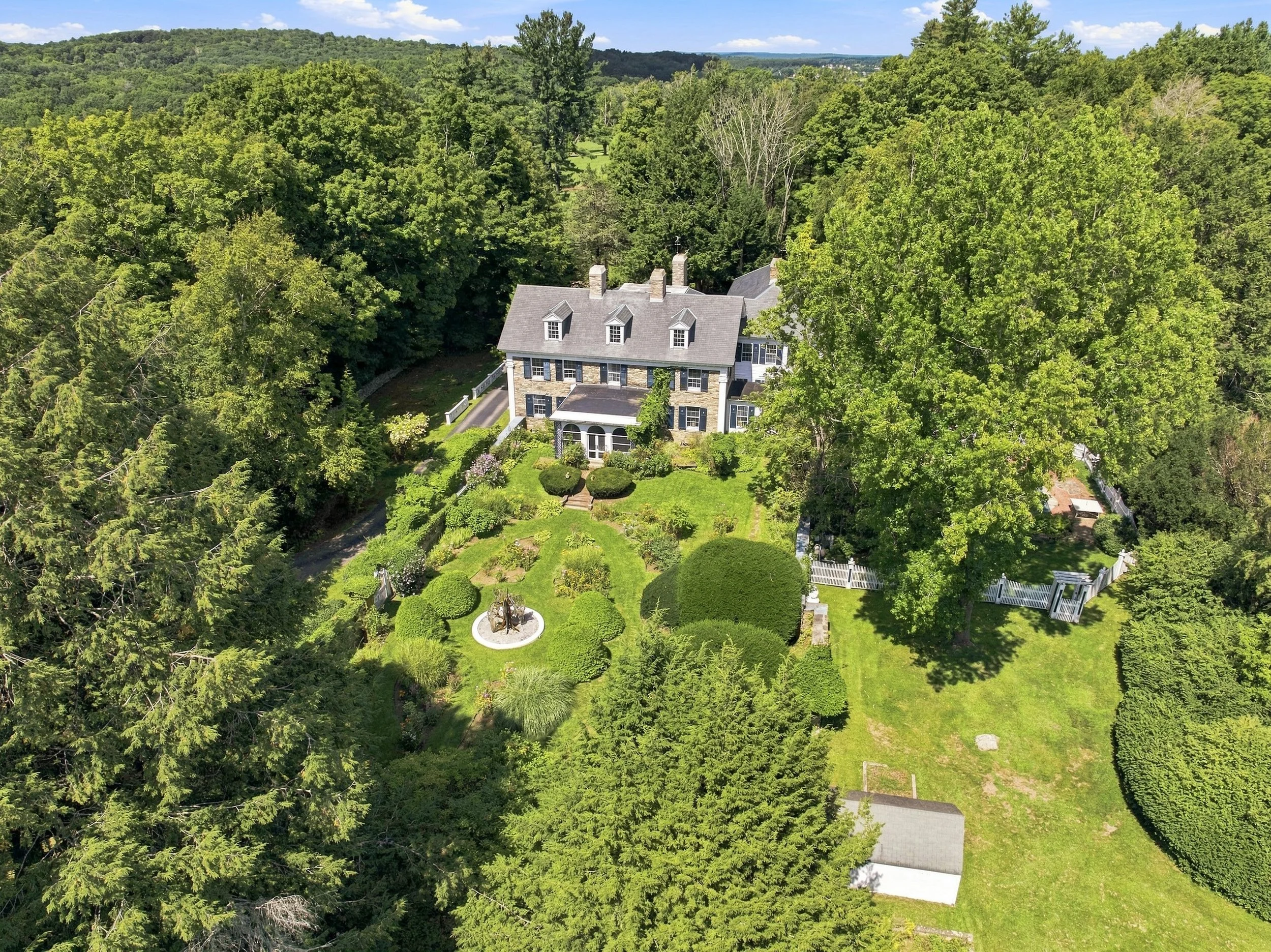A large house with a gray roof and stone exterior is surrounded by lush green trees and a well-maintained garden, situated in a rural area with rolling hills in the background.