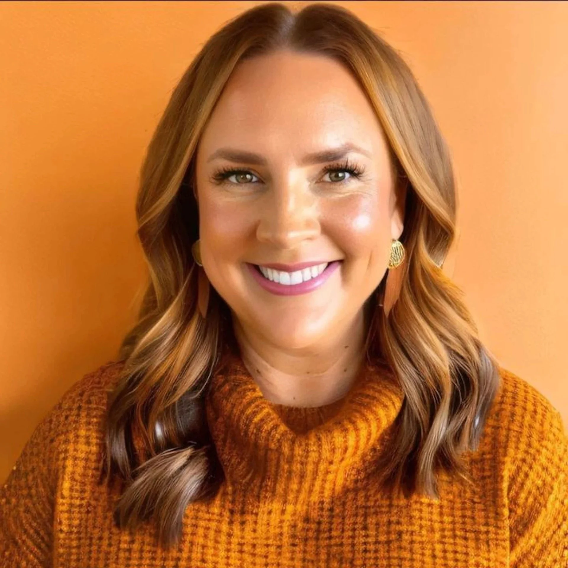 A woman with red hair, green eyes, and earrings, smiling, wearing an orange turtleneck sweater, in front of an orange background.