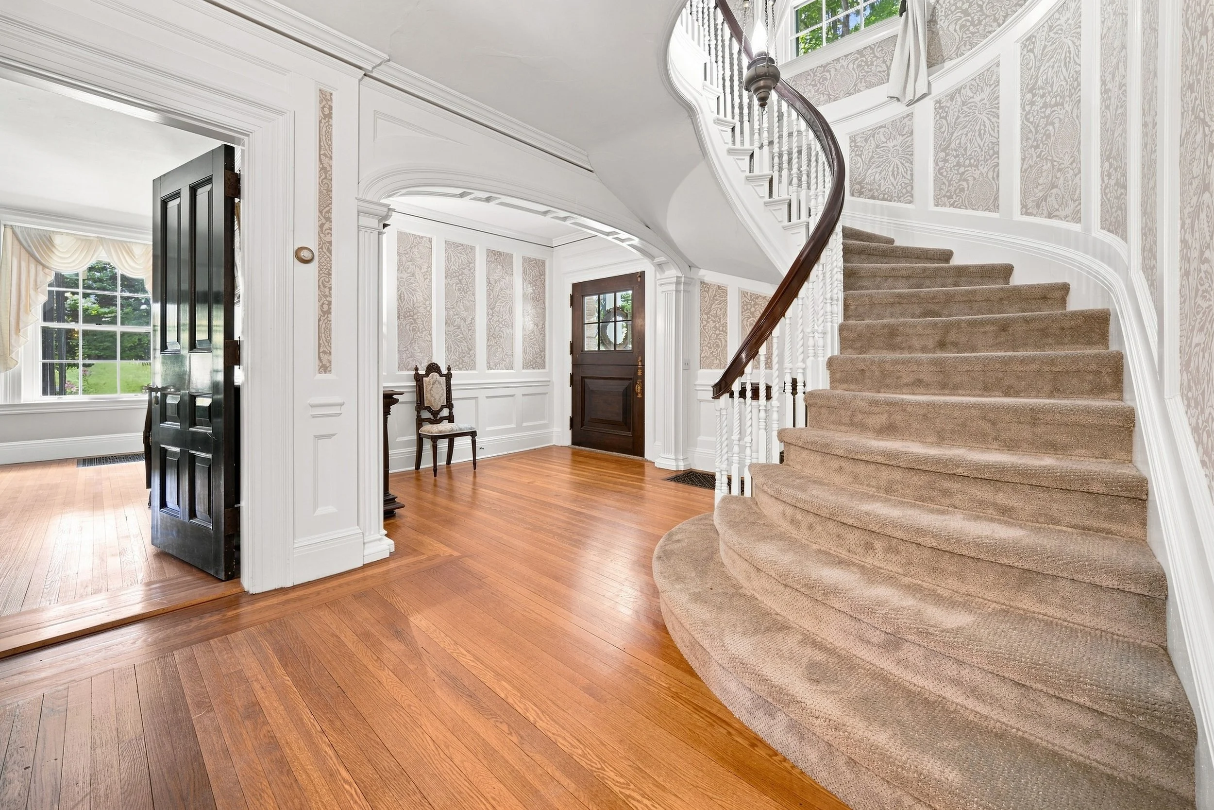 Interior of a house with a staircase, hardwood floors, a window with drapes, and an open door. The staircase has a wooden handrail and beige carpeted steps. Decorative wall paneling and wallpaper are visible.