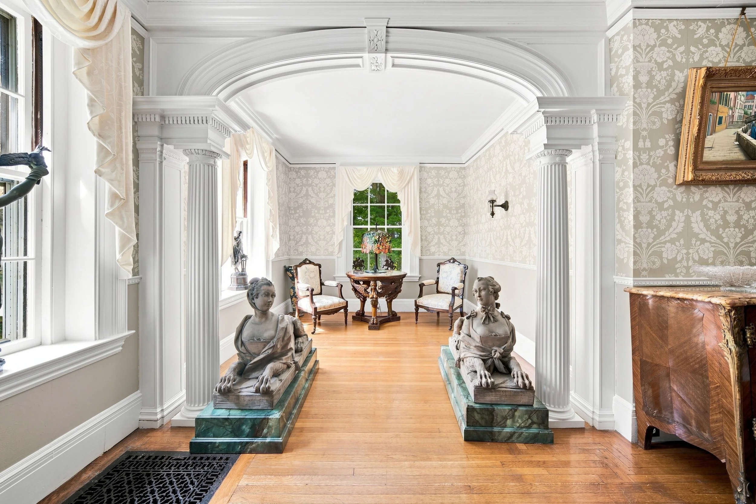 A spacious, elegant room with white decorative crown molding and columns, featuring two marble statues of seated women on pedestals. There are two chairs with ornate wooden frames and upholstery near a window, a small round table with a colorful flor