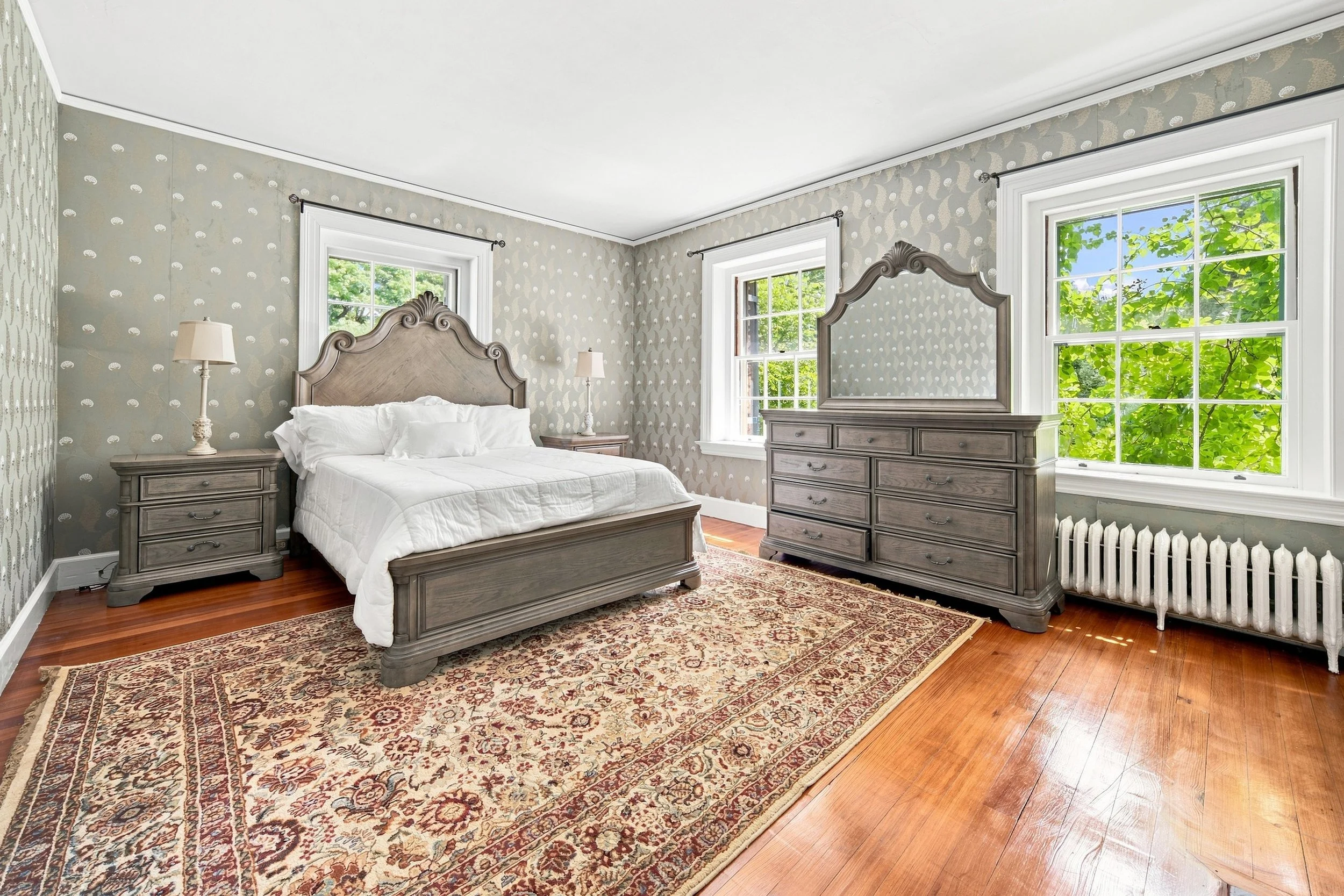 A bedroom with a bed, two nightstands with lamps, a dresser with a mirror, windows showing trees outside, an area rug, and hardwood floors.