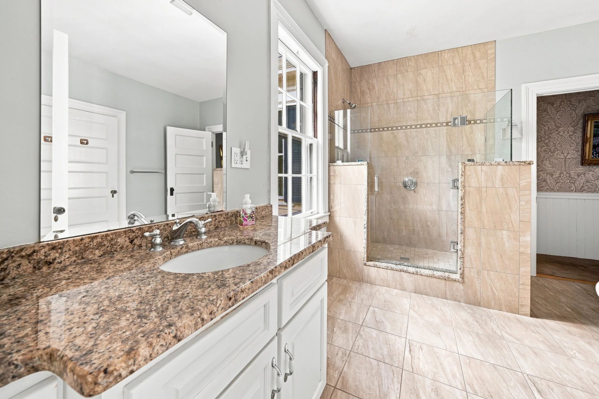A bathroom with a beige tiled shower area and a granite countertop vanity with a sink, large mirror, and soap dispensers.