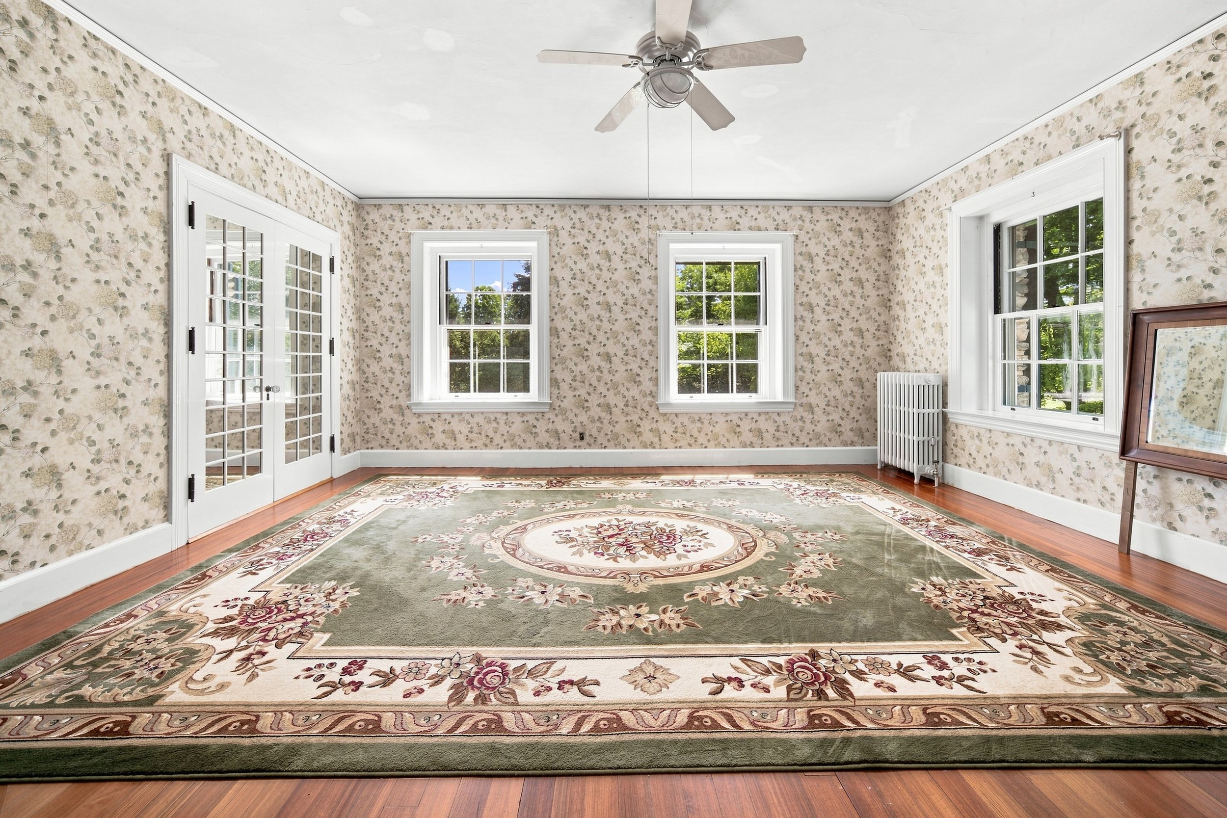 Empty living room with floral wallpaper, three windows showing green trees outside, white blinds, a ceiling fan, hardwood floors, a patterned area rug, a white radiator, and a framed picture leaning against the wall.