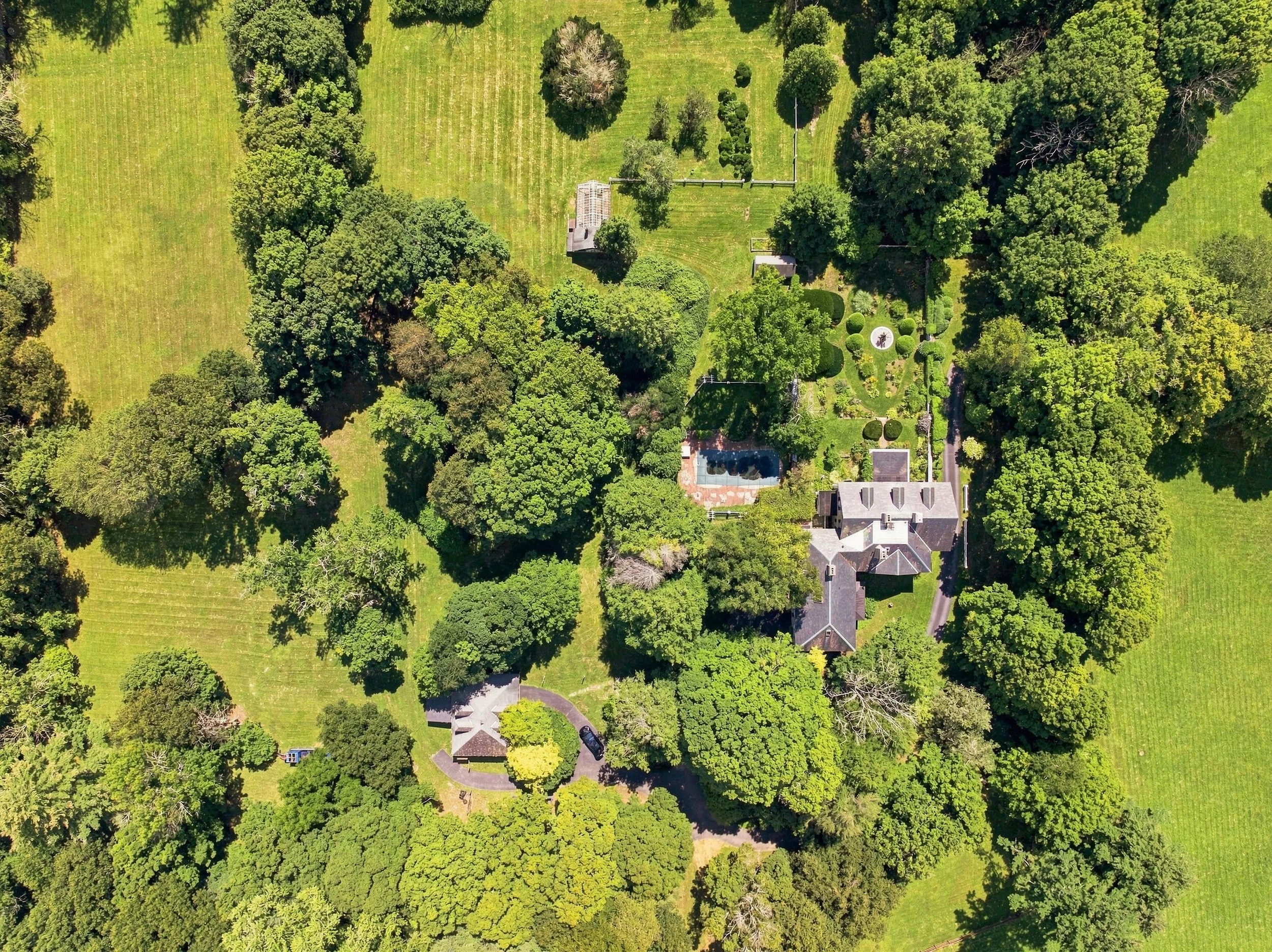 An aerial view of a lush green property with various trees, a house, a swimming pool, and well-maintained lawns