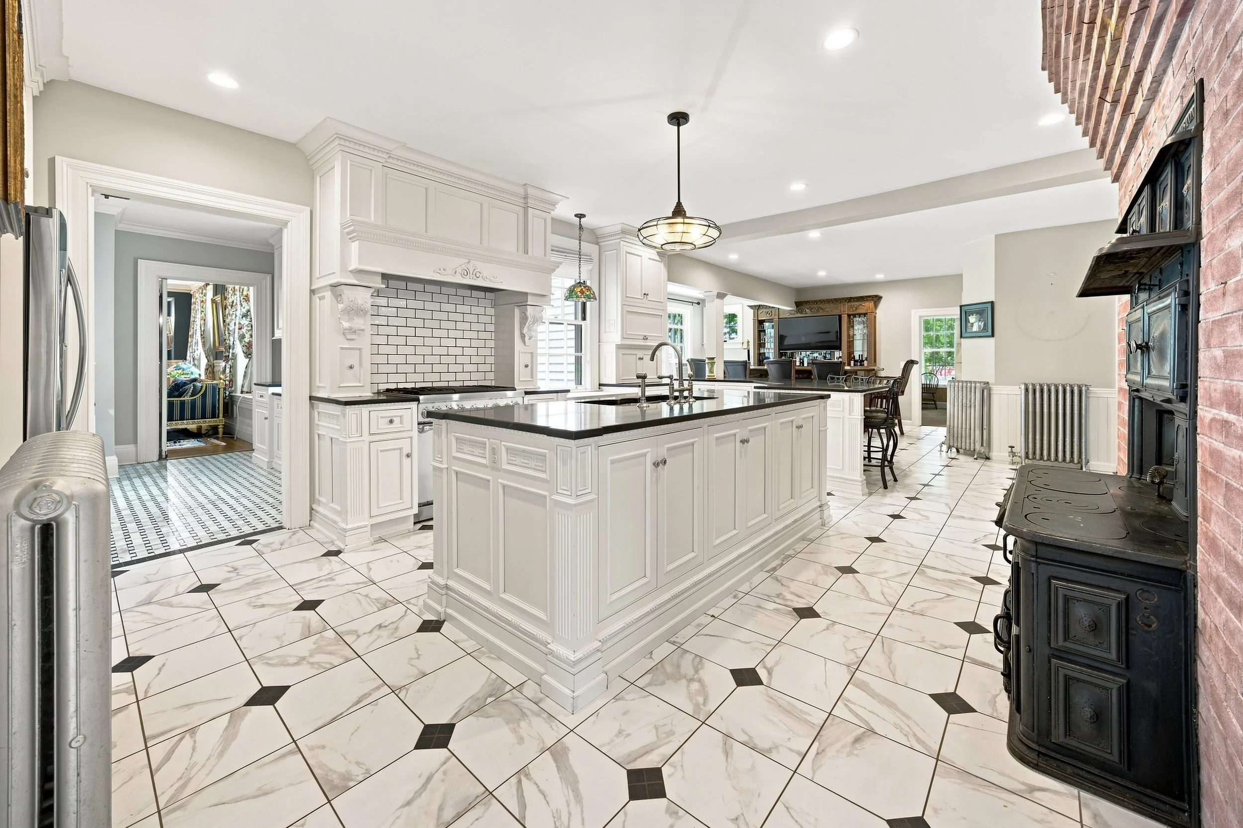Bright kitchen with white cabinetry, black countertops, tiled floor with black diamond accents, and a brick accent wall with a black stove. There's a large island in the center with a sink, and a dining area with a television in the background.